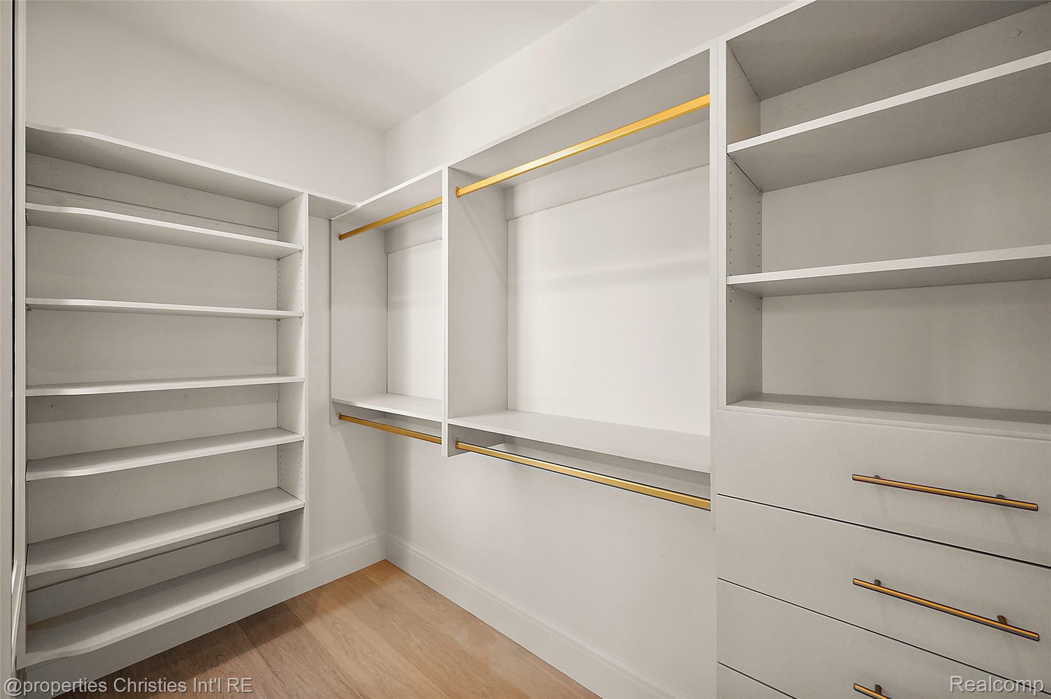 Walk-in closet with white built-in shelving, gold-tone hanging rods, drawers, and wood flooring.