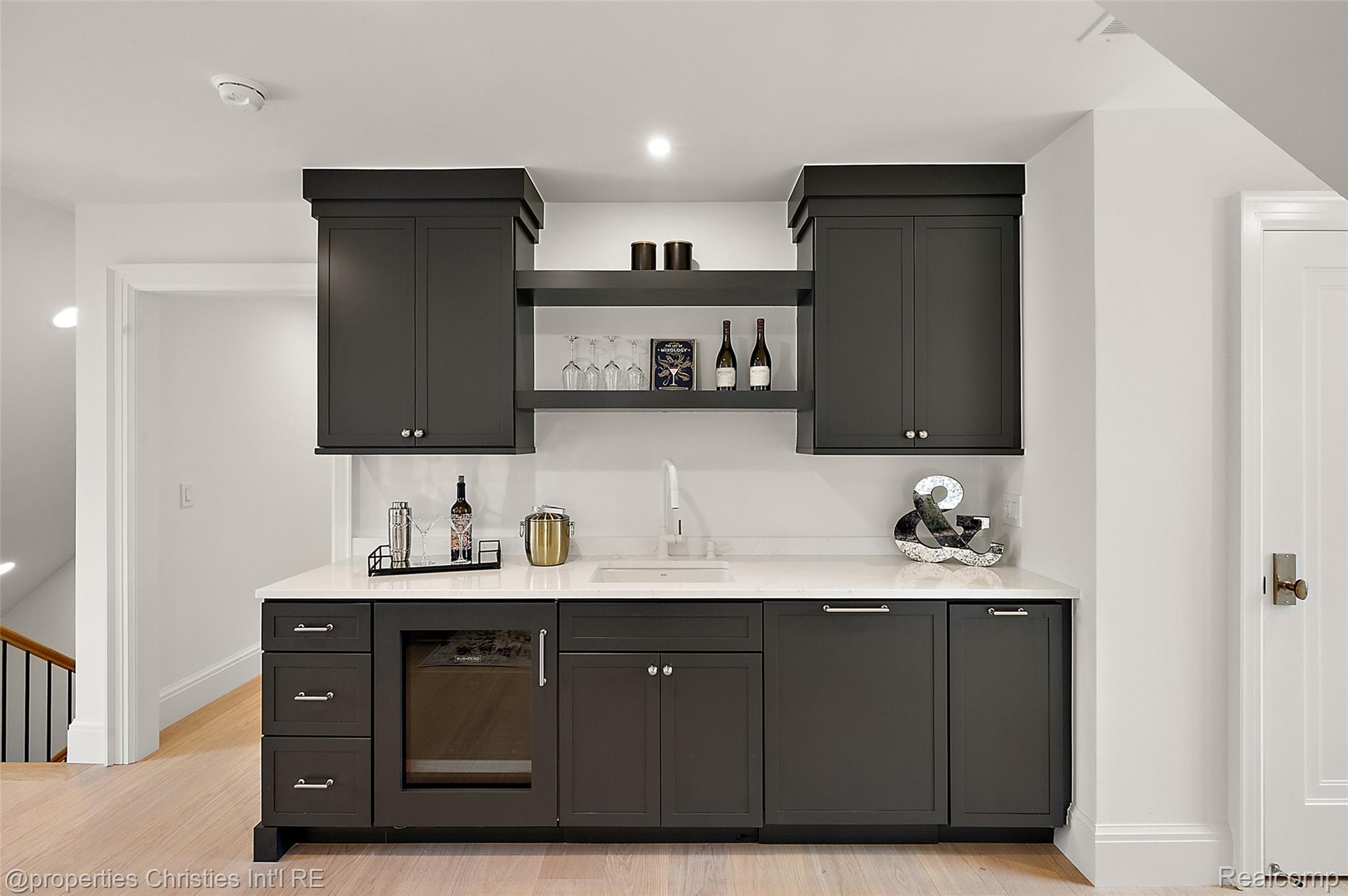 Wet bar with charcoal cabinetry, white countertop, open shelves, and an under-counter wine refrigerator.