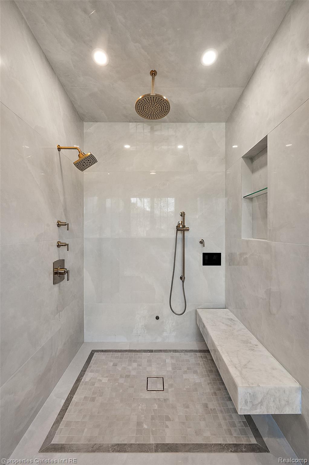 Walk-in shower with brass fixtures, pale stone tile, a built-in bench, and a recessed shelf.