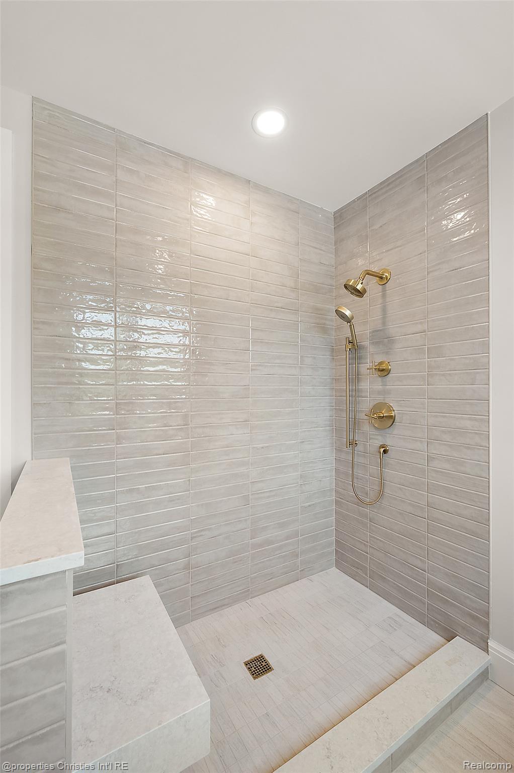 Walk-in shower with glossy gray tile, a stone bench, and brass rainfall and handheld fixtures.