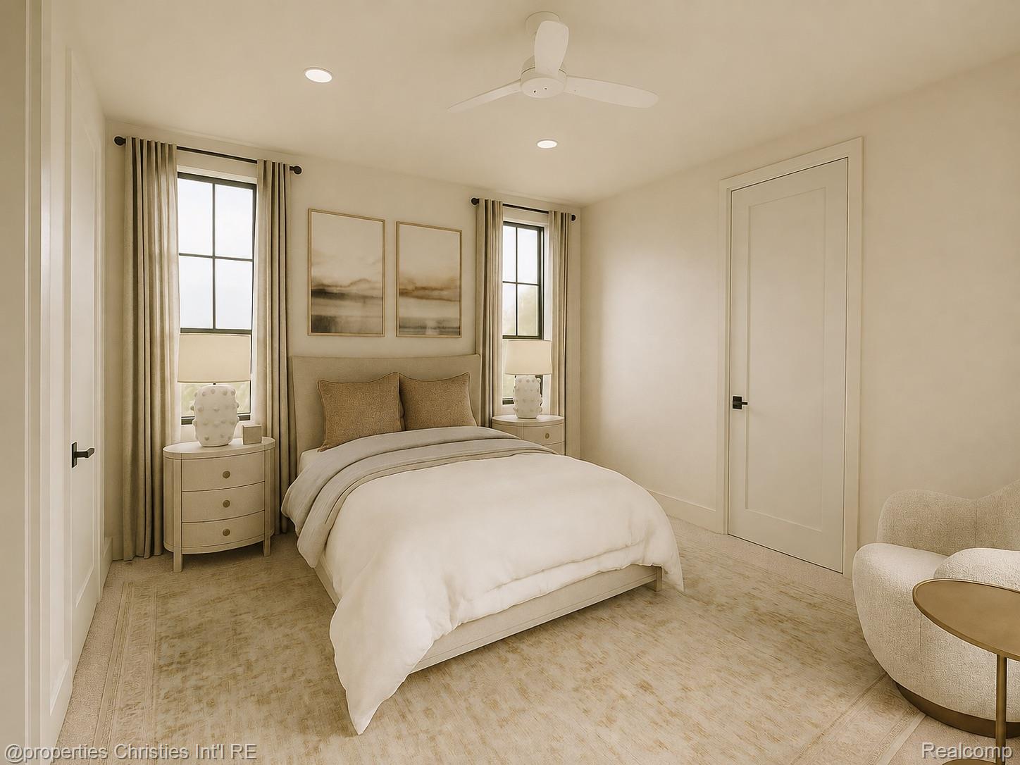 Bedroom with a cream bed between two tall curtained windows and a small seating area.