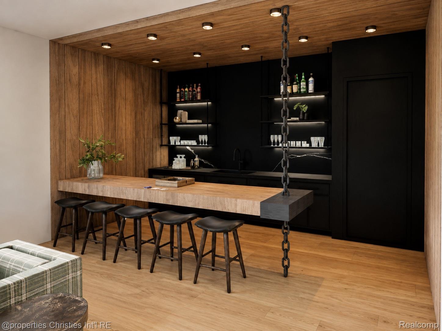 Modern home bar with a long wood island, black stools, and illuminated glassware shelves.