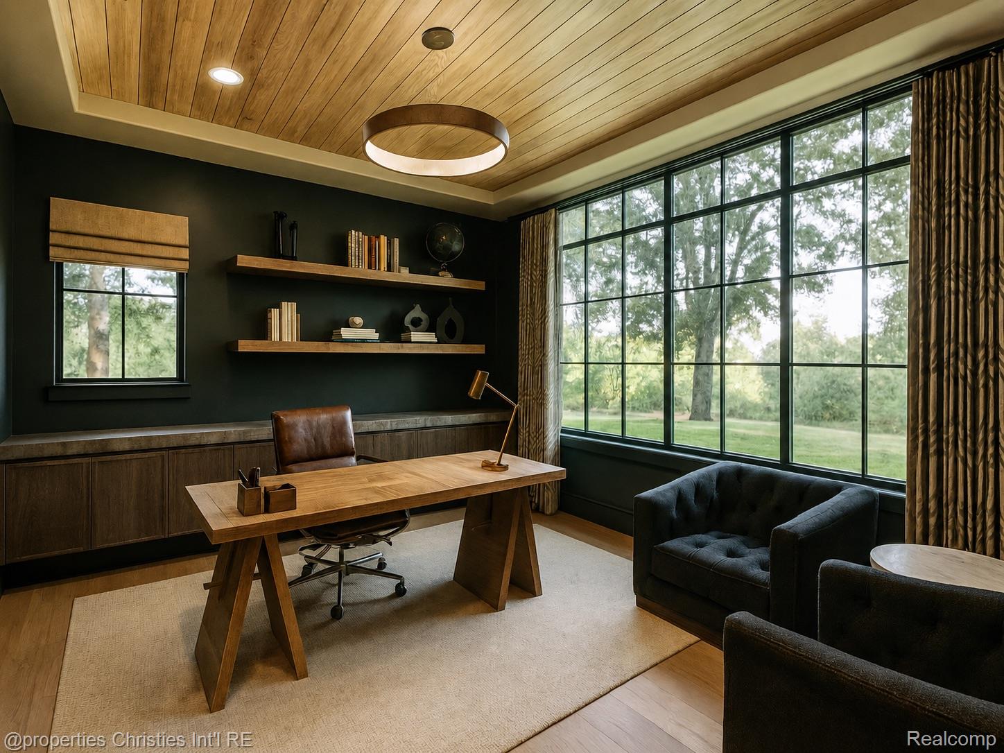 Furnished home office with a large wood desk, plank ceiling, and tall windows overlooking trees.