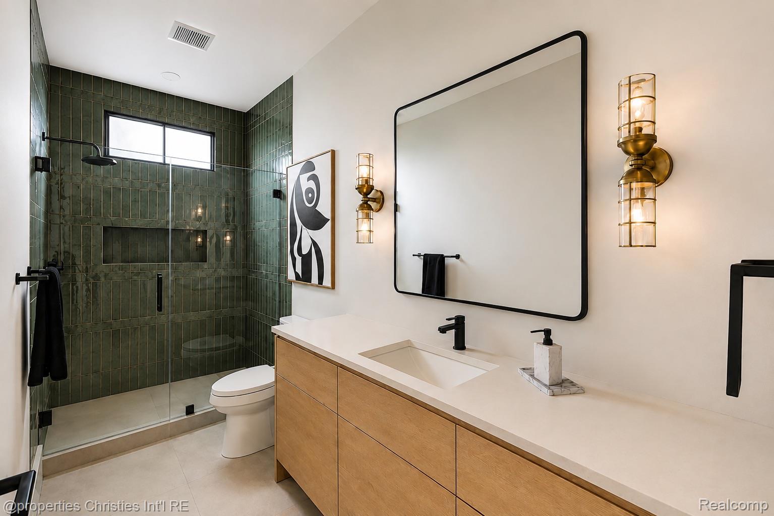 Modern bathroom with a green-tiled glass shower, wood vanity, large mirror, and brass sconces.