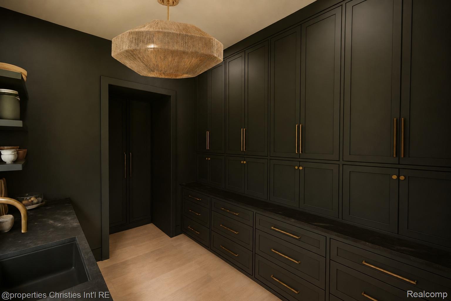 Dark paneled kitchen pantry with brass cabinet pulls, black stone counters, and a woven pendant light.