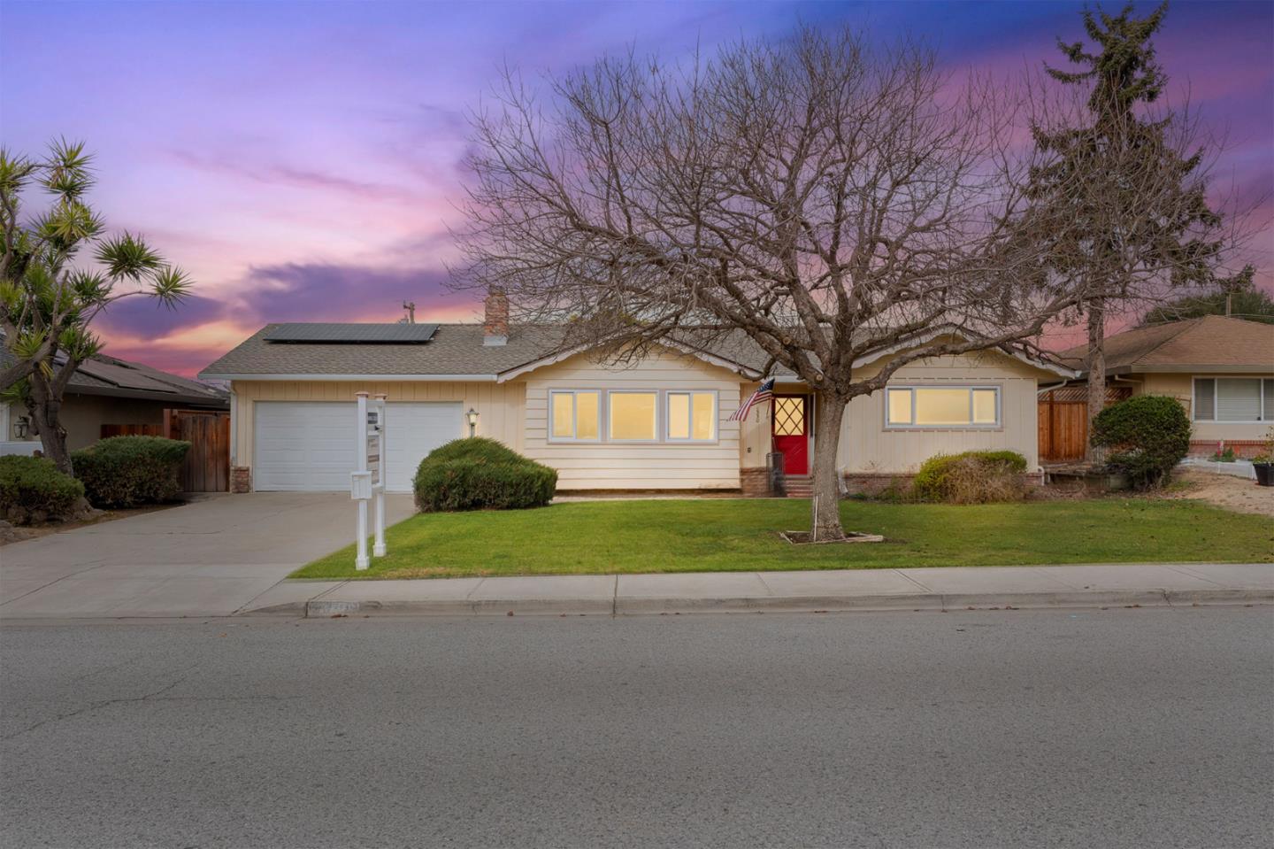 Single-story beige home with rooftop solar panels and a red front door beneath a large leafless tree.
