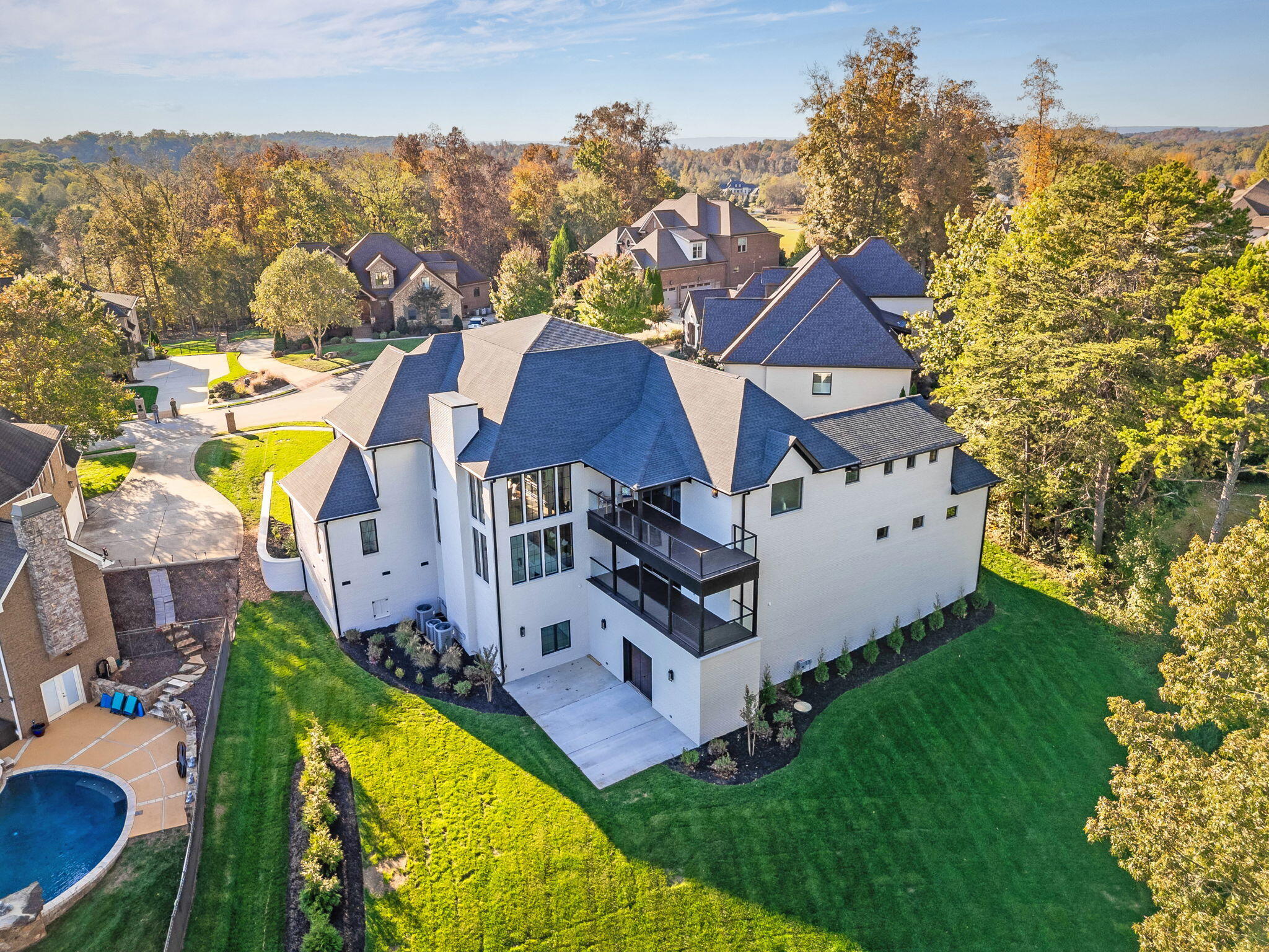 Aerial view of a white multi-story home with a dark roof, balcony, and broad lawn amid autumn trees.
