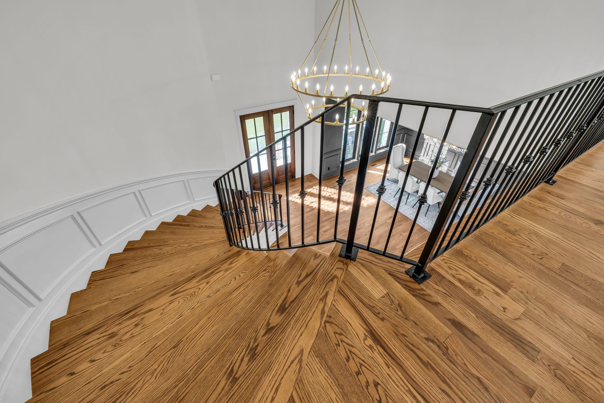 Curved oak staircase with black railings overlooking a dining area beneath a tiered gold chandelier.