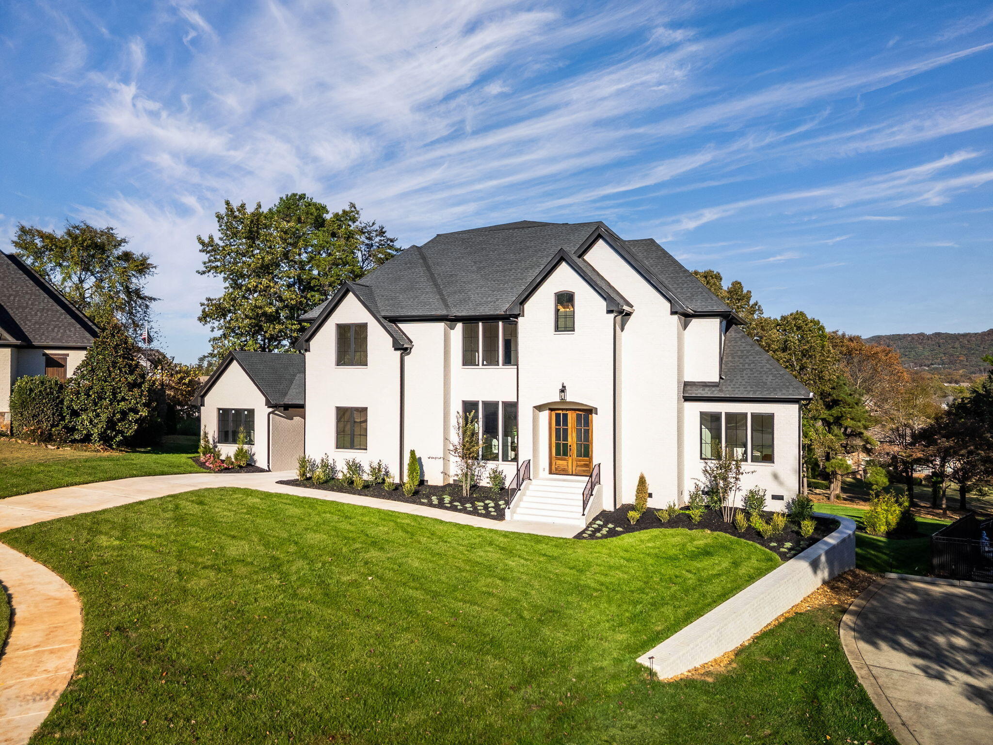 White two-story house with dark gabled roofs, wood double doors, and a landscaped lawn.