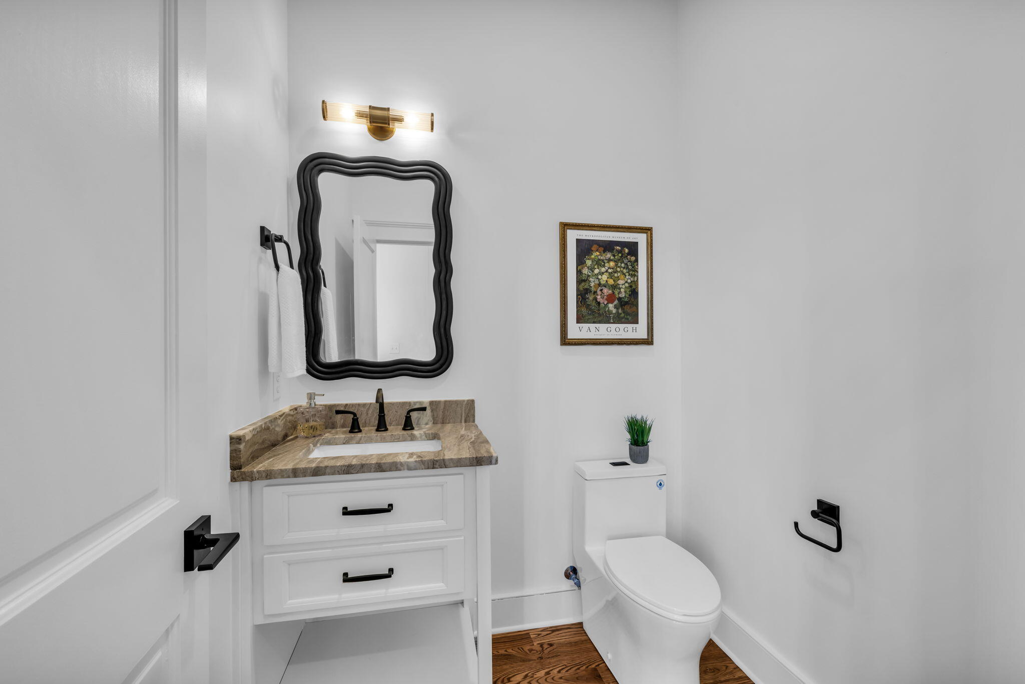 Bathroom with a stone-countertop vanity, wavy black mirror, toilet, and framed floral artwork.