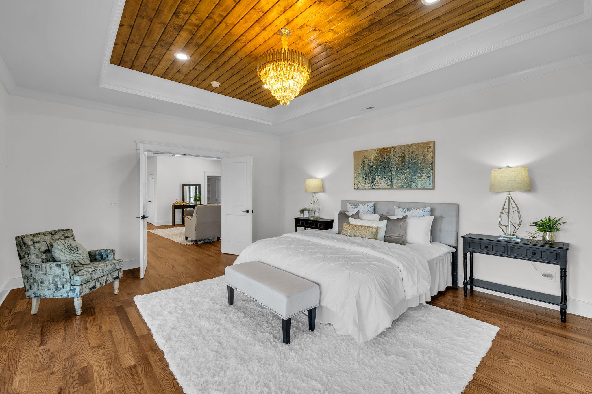 Staged bedroom with a white bed beneath a wood-plank ceiling and gold chandelier.