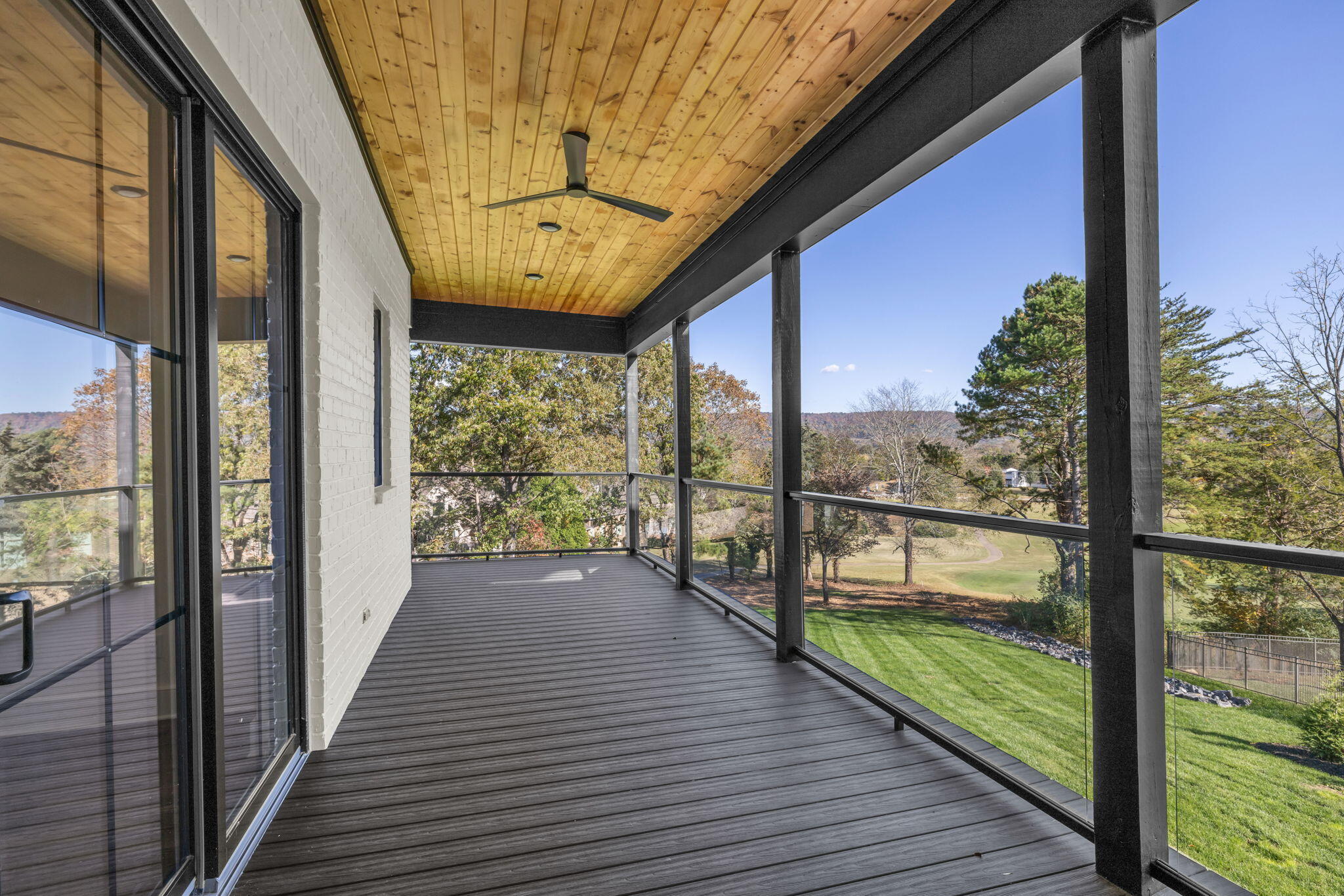 Covered deck with a wood ceiling and glass railings overlooking a lawn and wooded ridge.