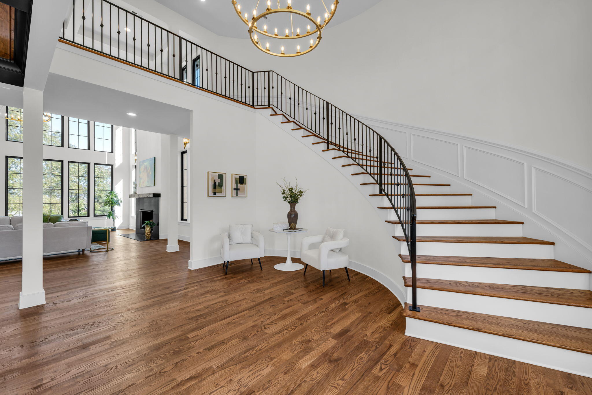 Bright foyer with curved wood staircase, brass chandelier, hardwood floors, and tall windows.