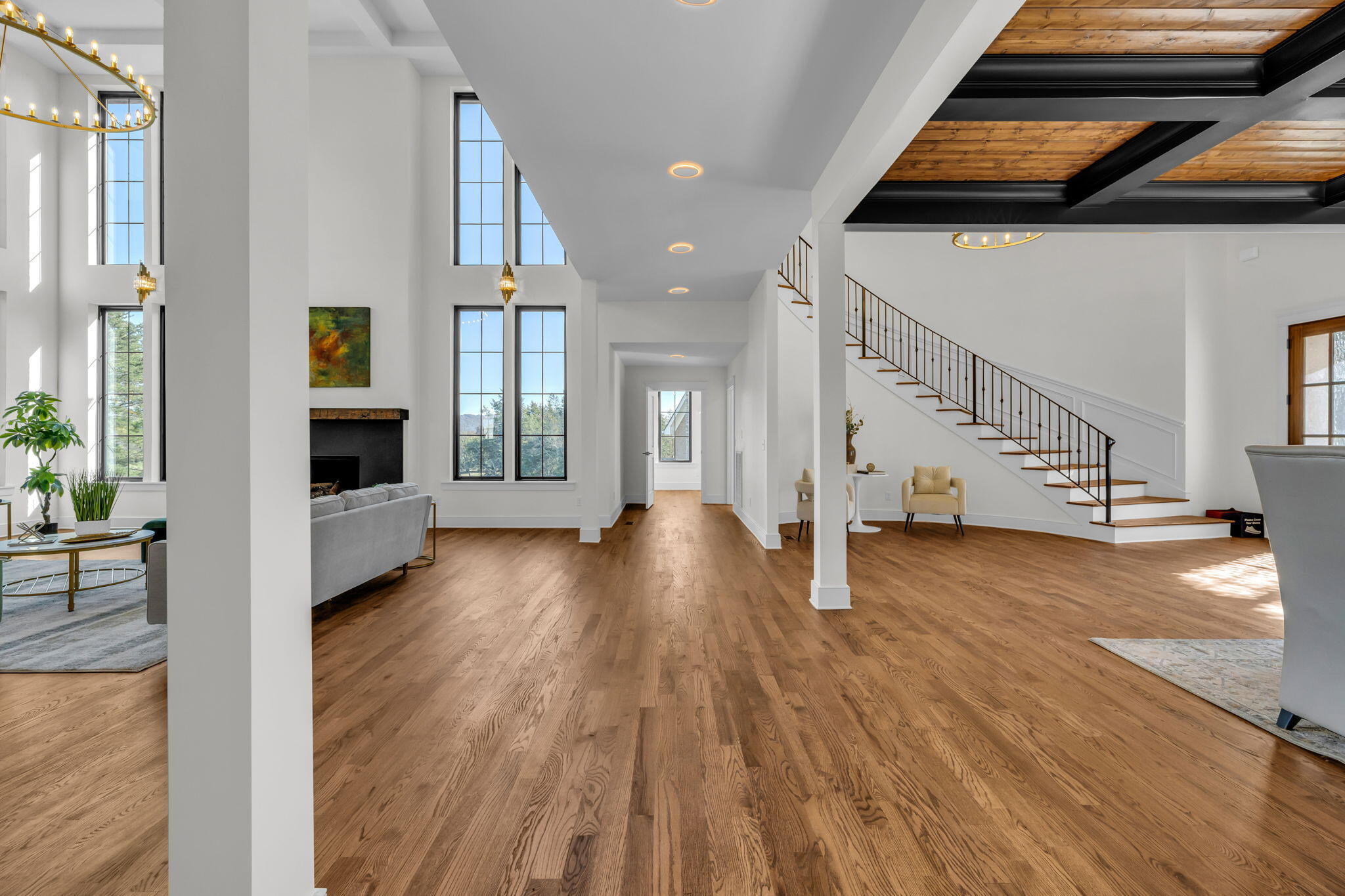 Open-plan living room with tall windows, hardwood floors, a fireplace, and a staircase beneath a wood ceiling.
