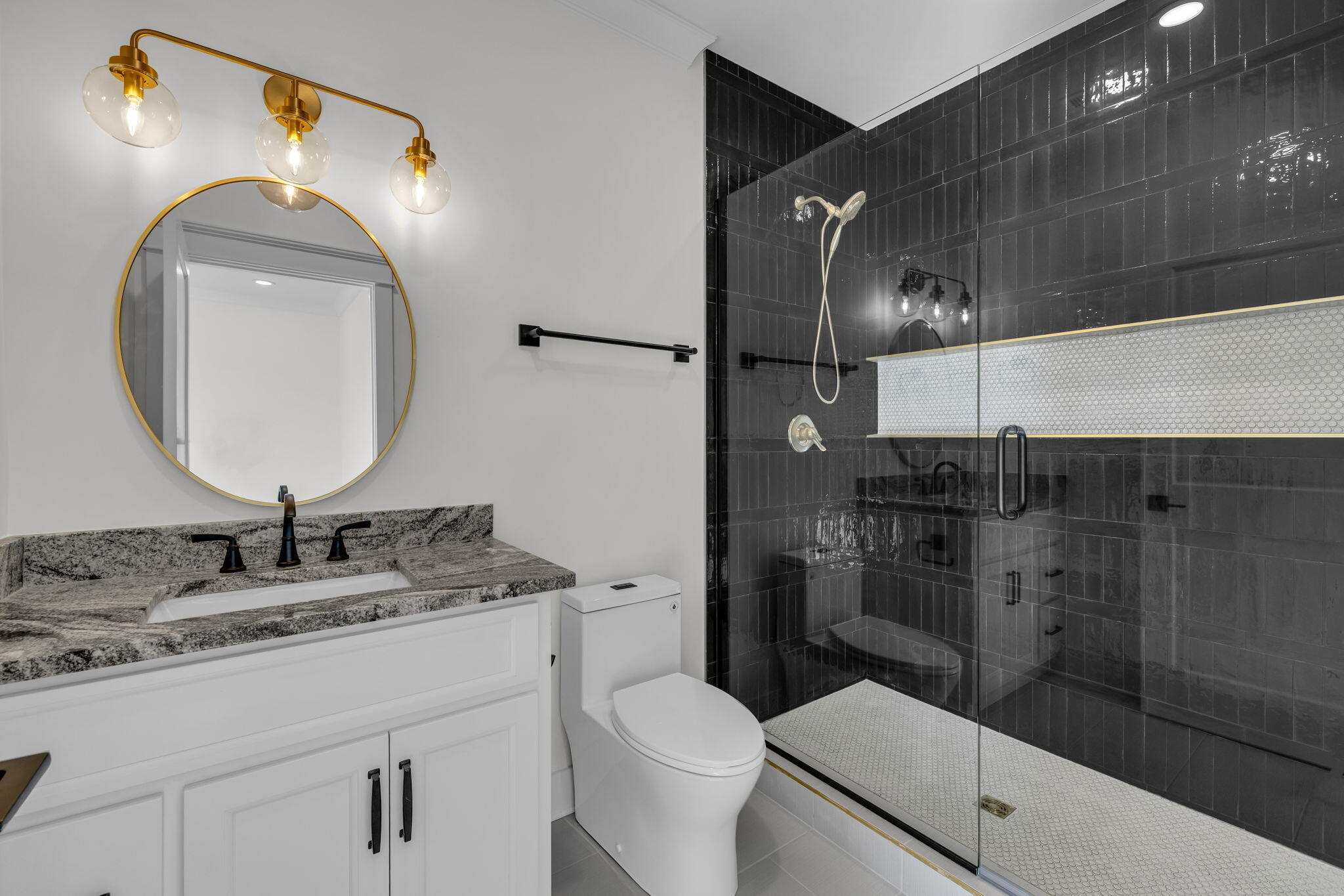 Bathroom with a glass walk-in shower, dark tile, and white vanity beneath a round gold-framed mirror.