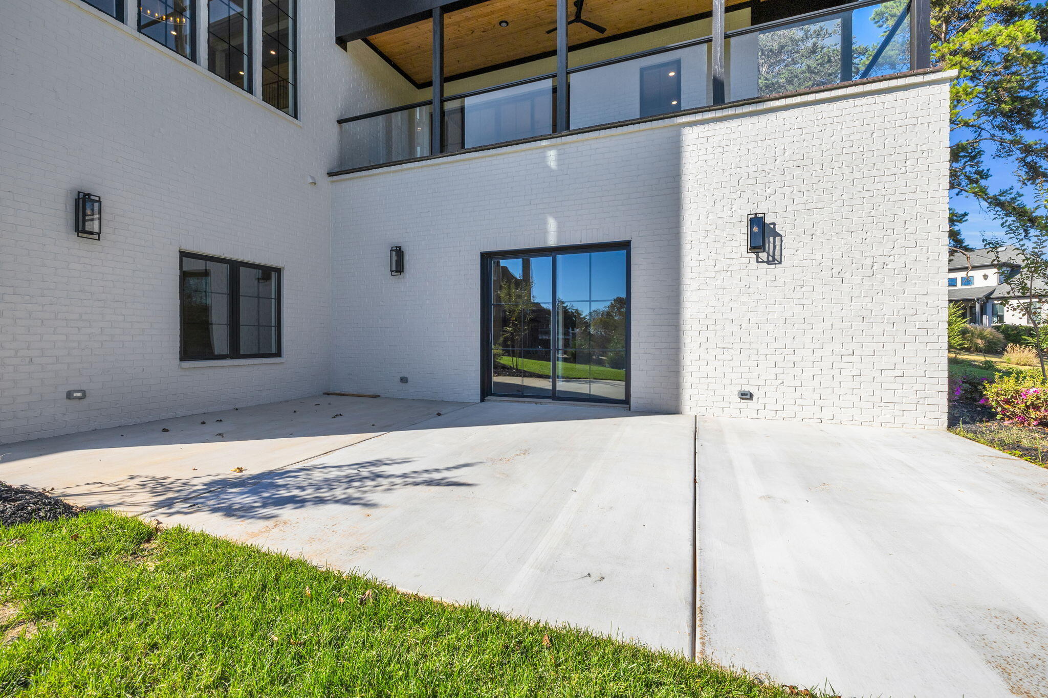 Modern white-brick home with a broad concrete patio, sliding glass doors, and an upper-level balcony.