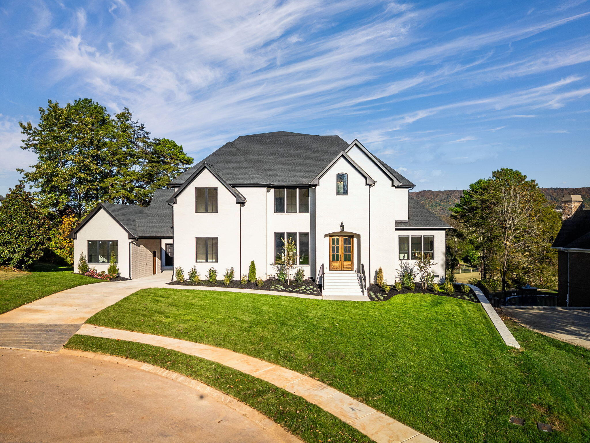 White two-story house with dark gabled roofs, wood front door, landscaped lawn, and wooded hillside.