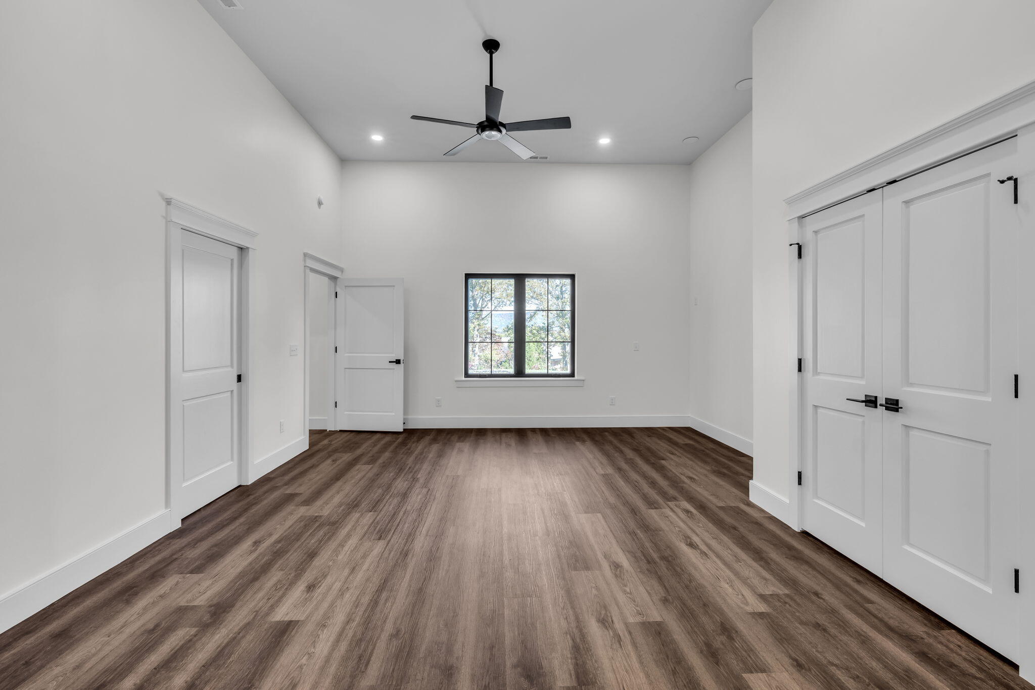 Empty room with wood-look flooring, a black ceiling fan, a window, and white paneled doors.
