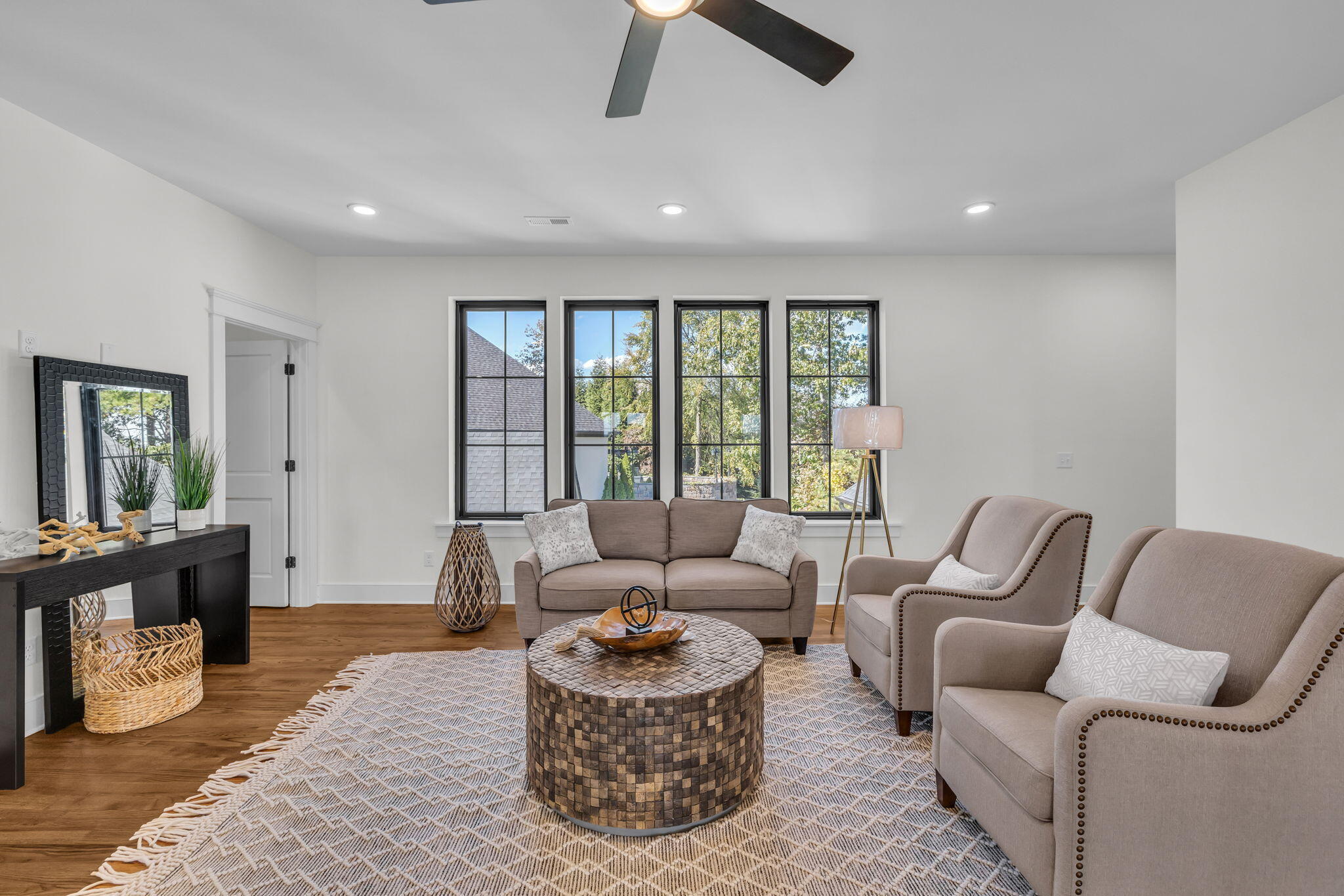 Staged living room with taupe seating, a round wood coffee table, and black-framed windows.