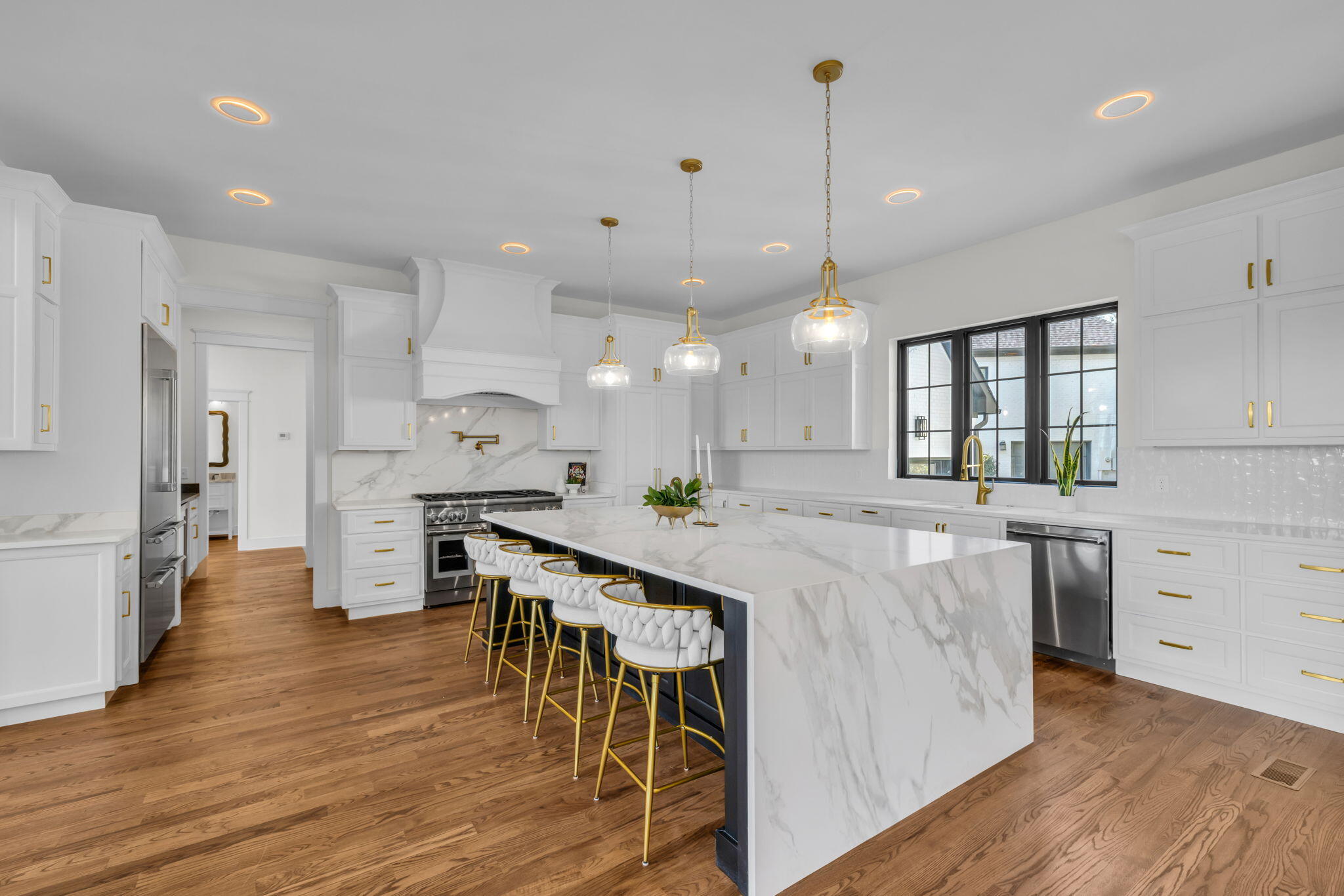 White kitchen with a large waterfall island, gold-accented bar stools, and hardwood floors.
