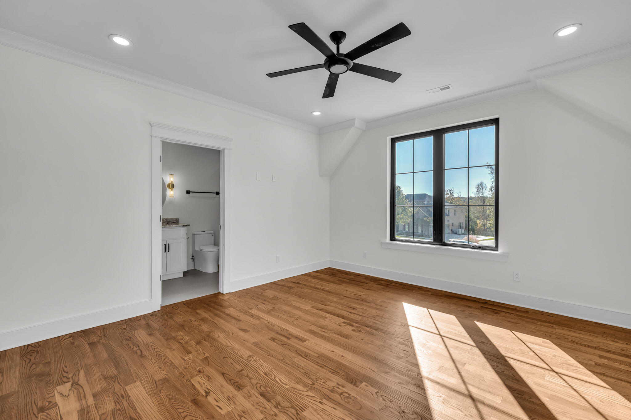 Empty room with hardwood floors, a black ceiling fan, and a large window beside an adjoining bathroom.