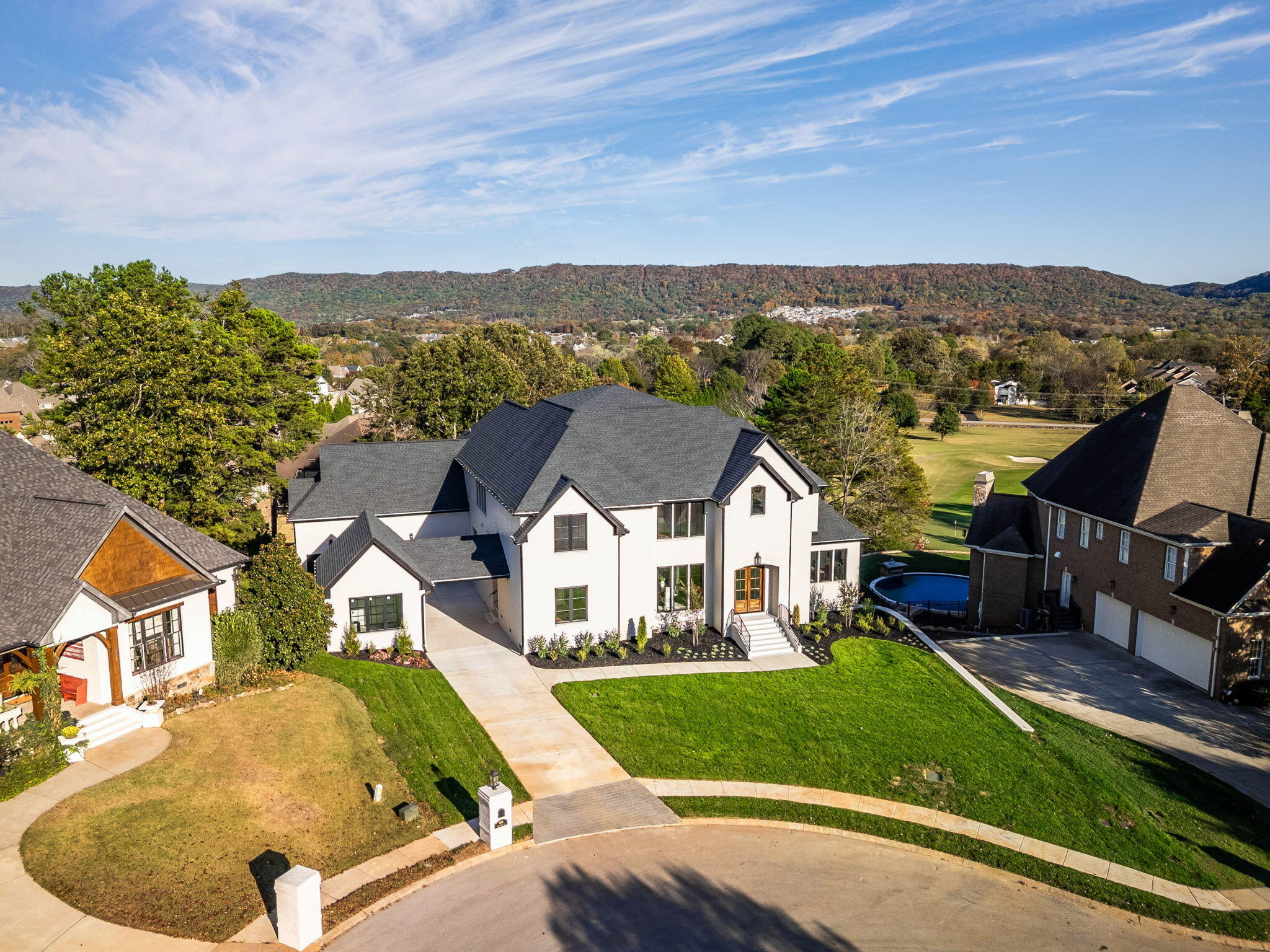 White two-story home with dark roof and broad driveway, viewed above the street with a golf course beyond.