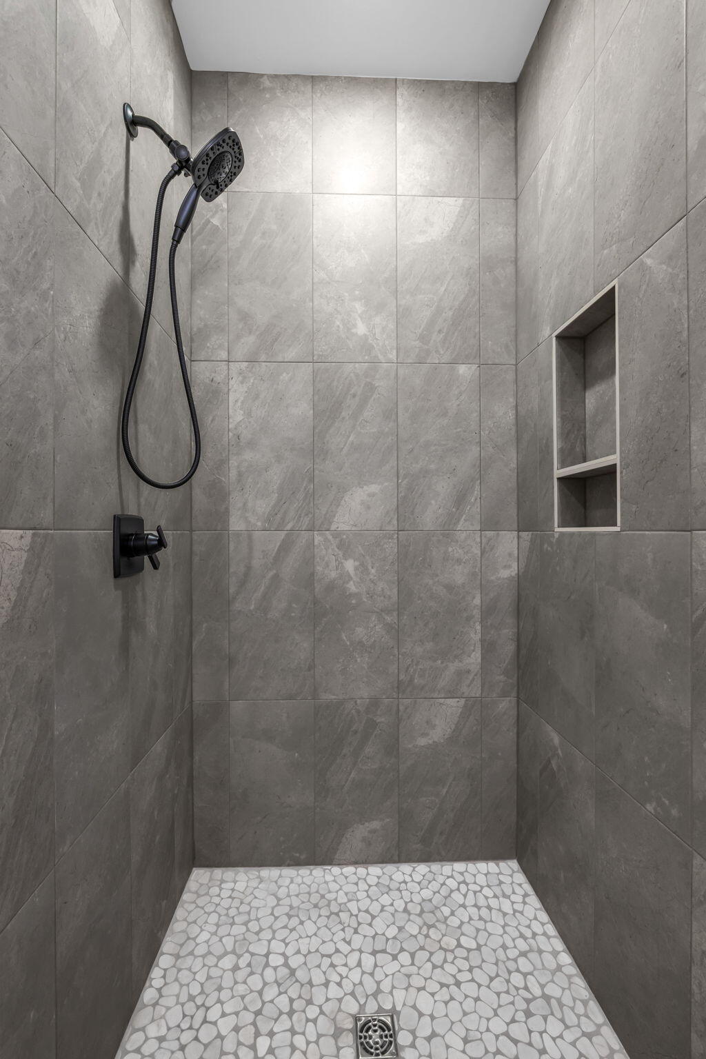 Walk-in shower with gray tile walls, a black handheld showerhead, recessed niche, and pebble-mosaic floor.
