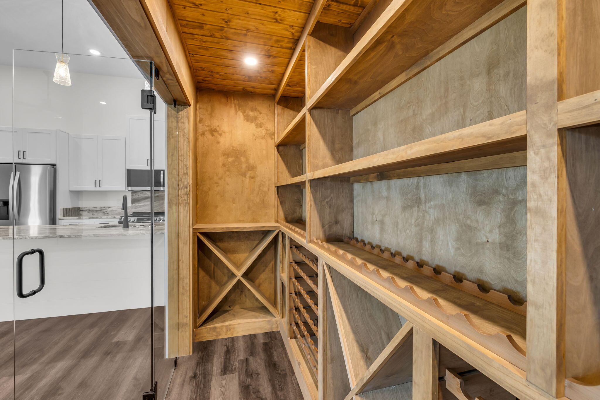 Wood wine cellar with built-in bottle racks and a glass door opening to a white kitchen.