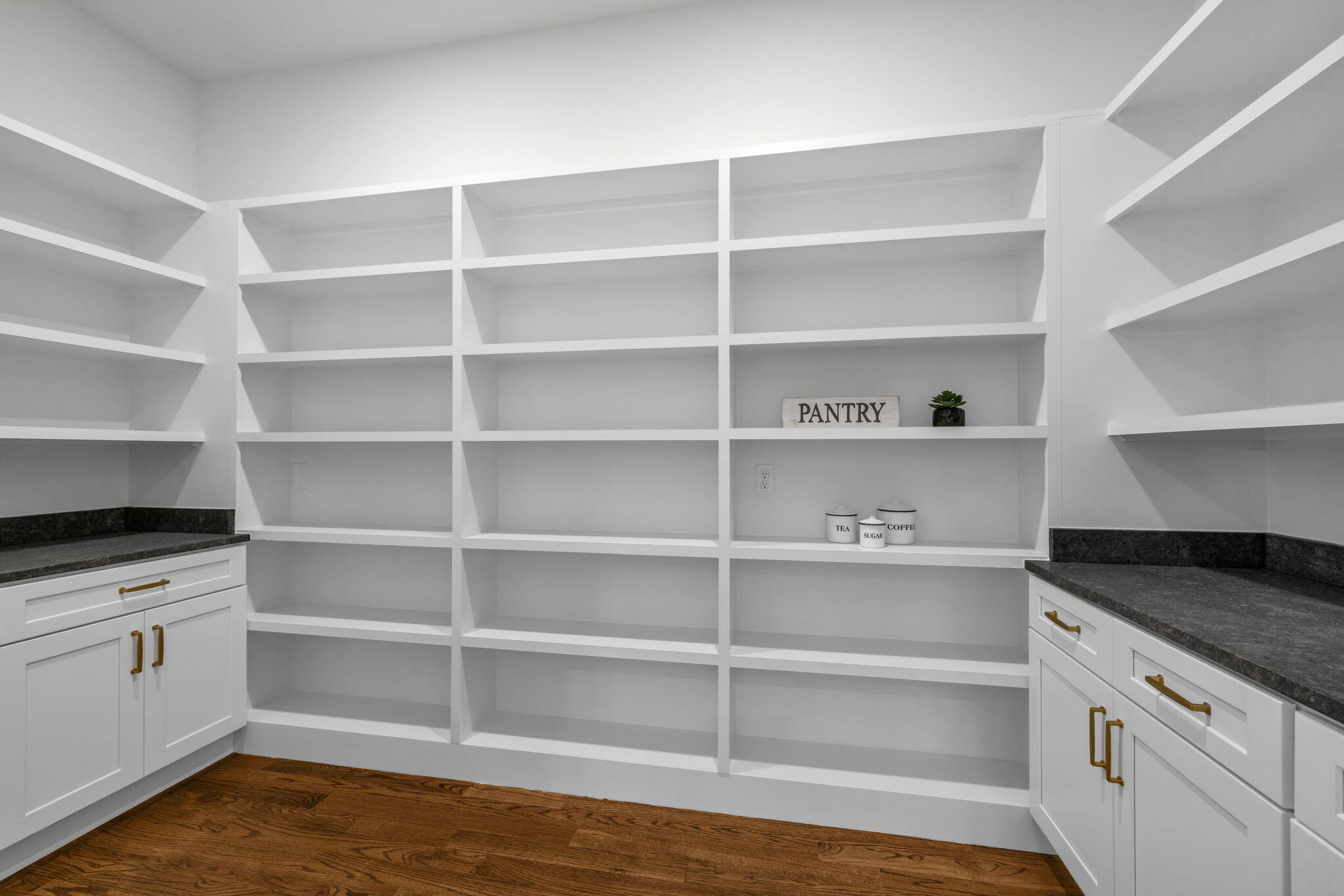 Walk-in pantry with white built-in shelves, dark countertops, brass cabinet pulls, and a wood floor.