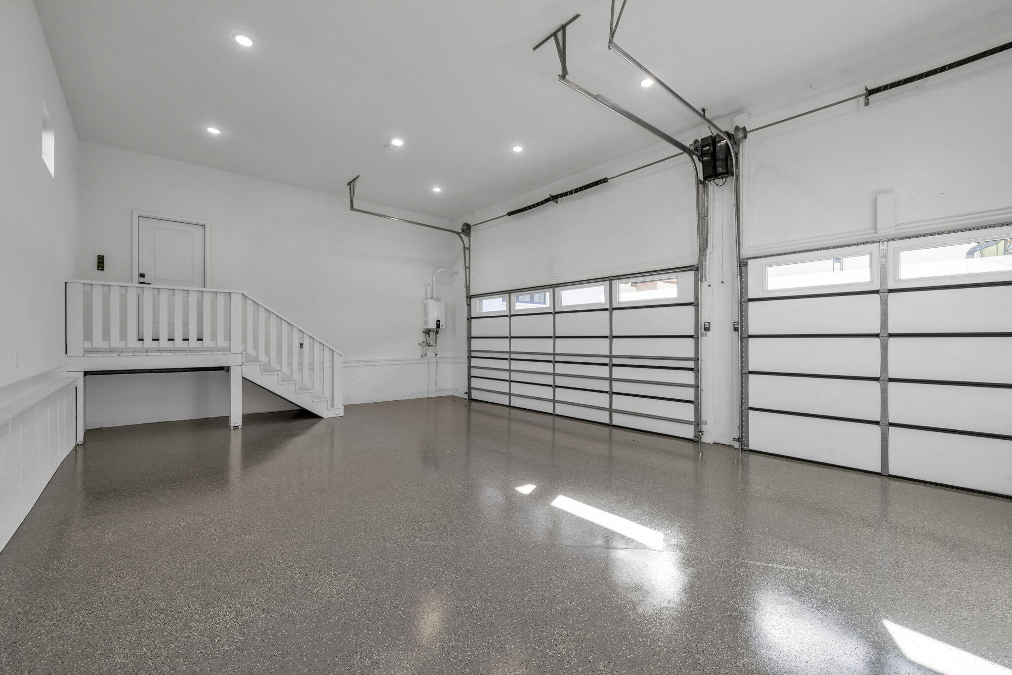 Vacant garage with two doors, a speckled epoxy floor, and stairs to a raised storage platform.