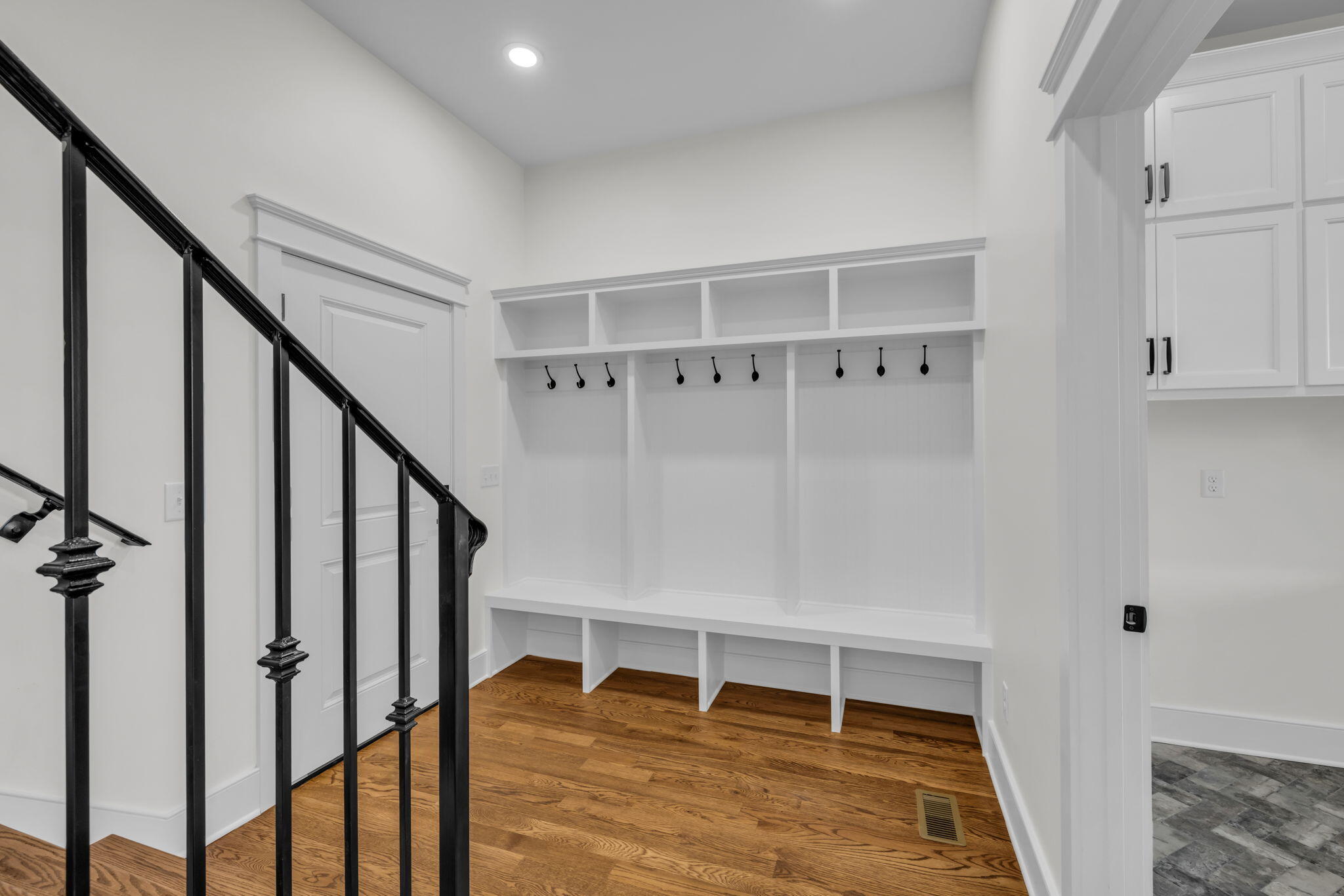 Mudroom with a built-in bench, cubbies, black hooks, hardwood flooring, and a black stair railing.