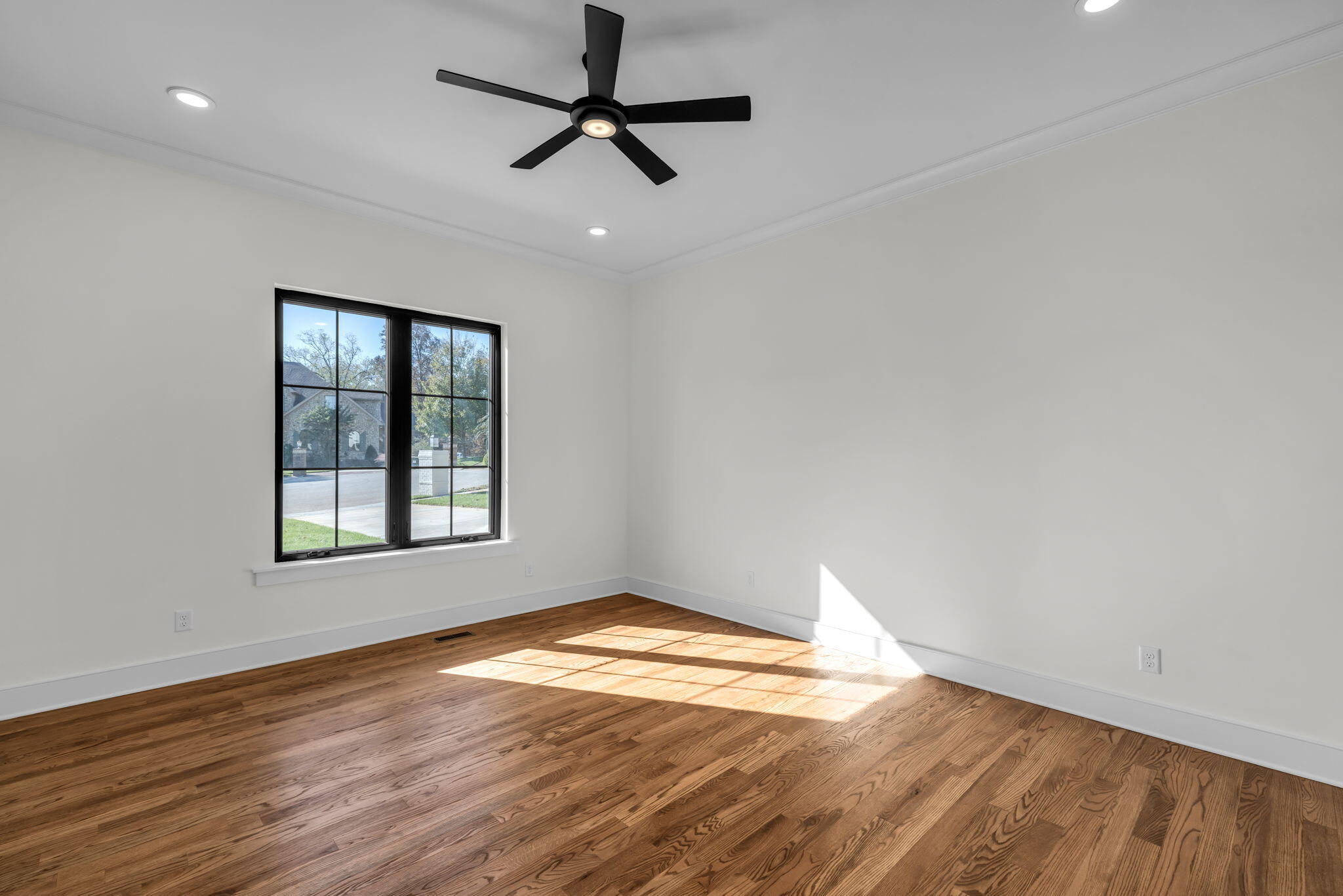 Empty room with hardwood floors, a black ceiling fan, and sunlight through a large window.