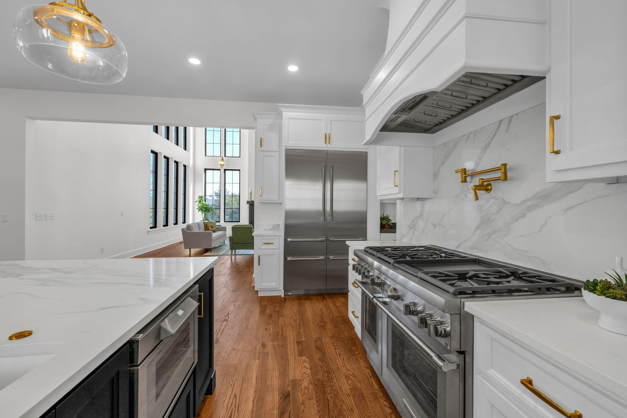 Modern white kitchen with a stainless-steel range, marble surfaces, gold hardware, and tall windows beyond.