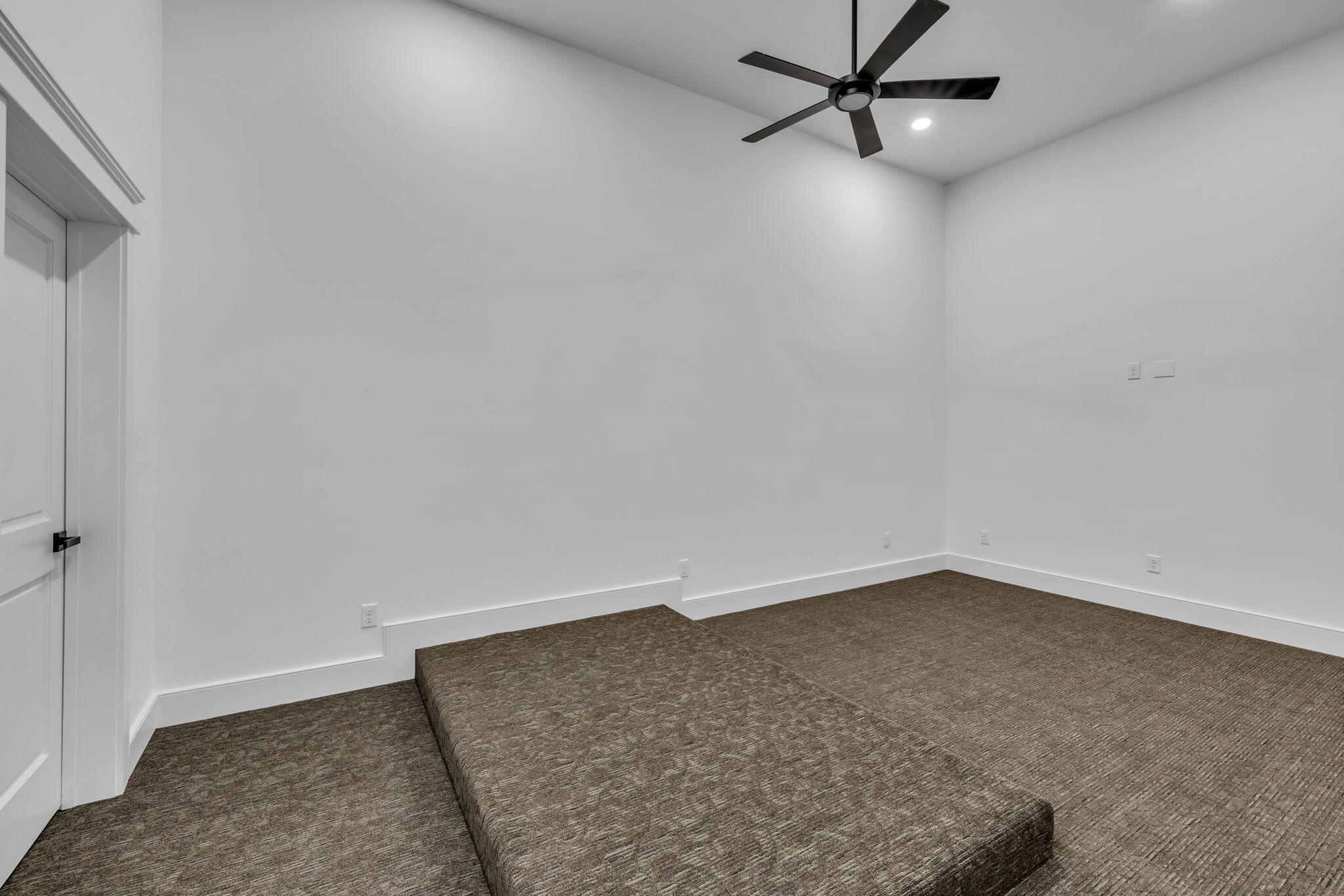 Empty carpeted room with a raised floor platform and dark ceiling fan.