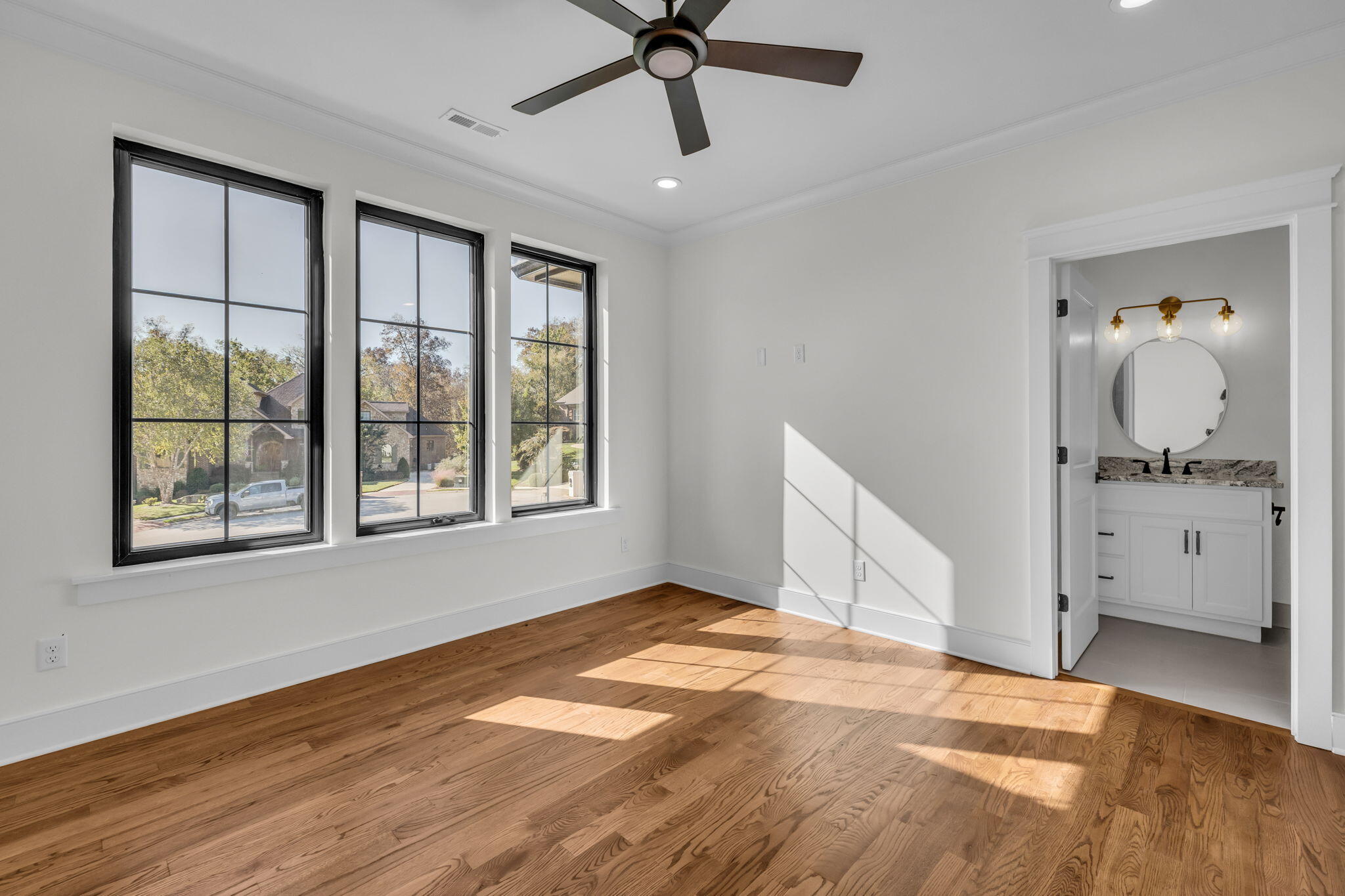 Empty bedroom with hardwood floors and tall windows, opening to a bathroom with a round mirror.