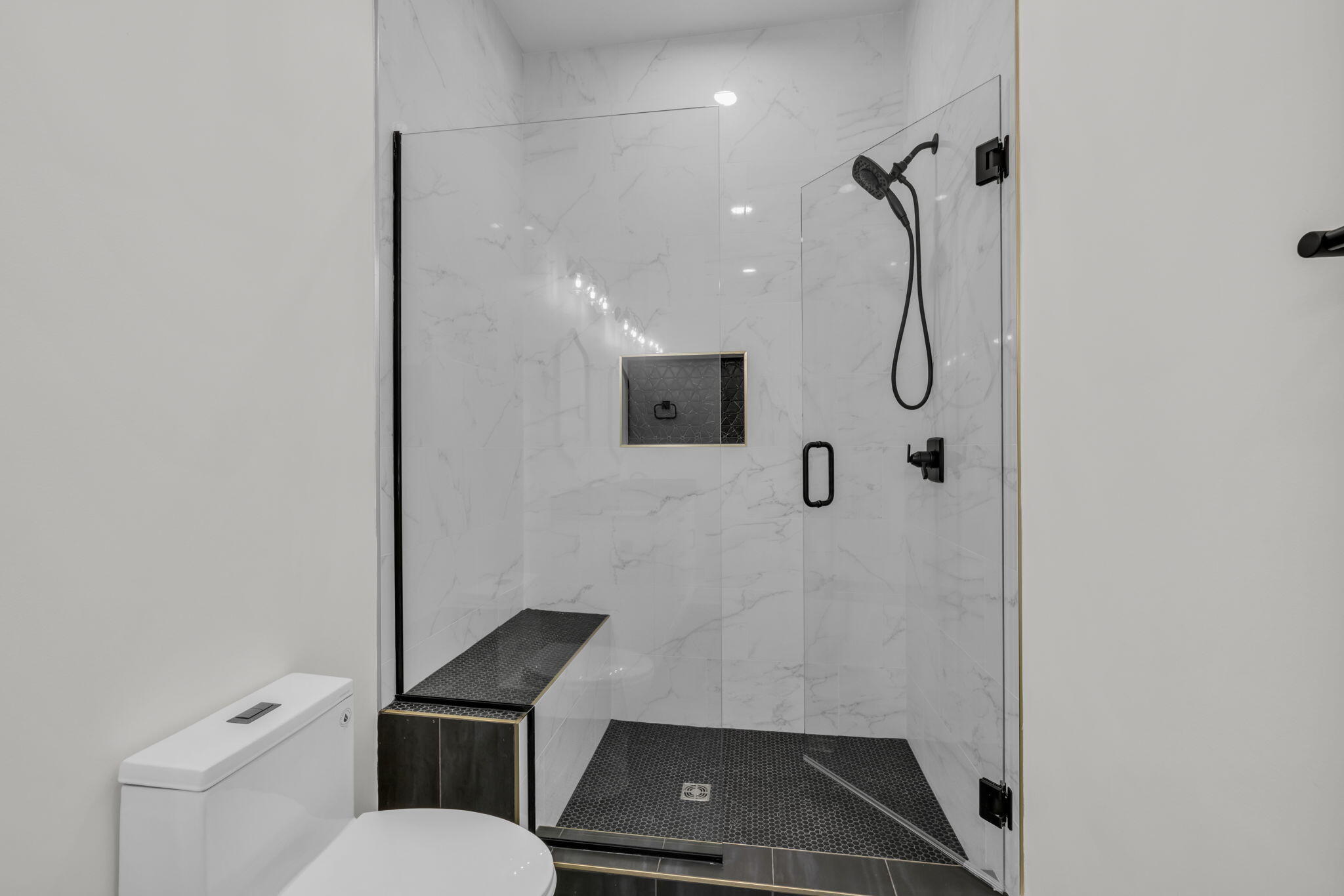 Walk-in shower with marble-look tile, black fixtures, mosaic floor, and built-in bench beside a toilet.
