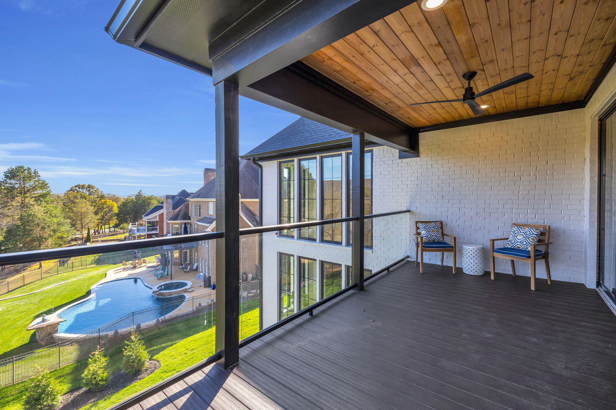 Covered balcony with a wood ceiling and glass railing overlooking a backyard pool.