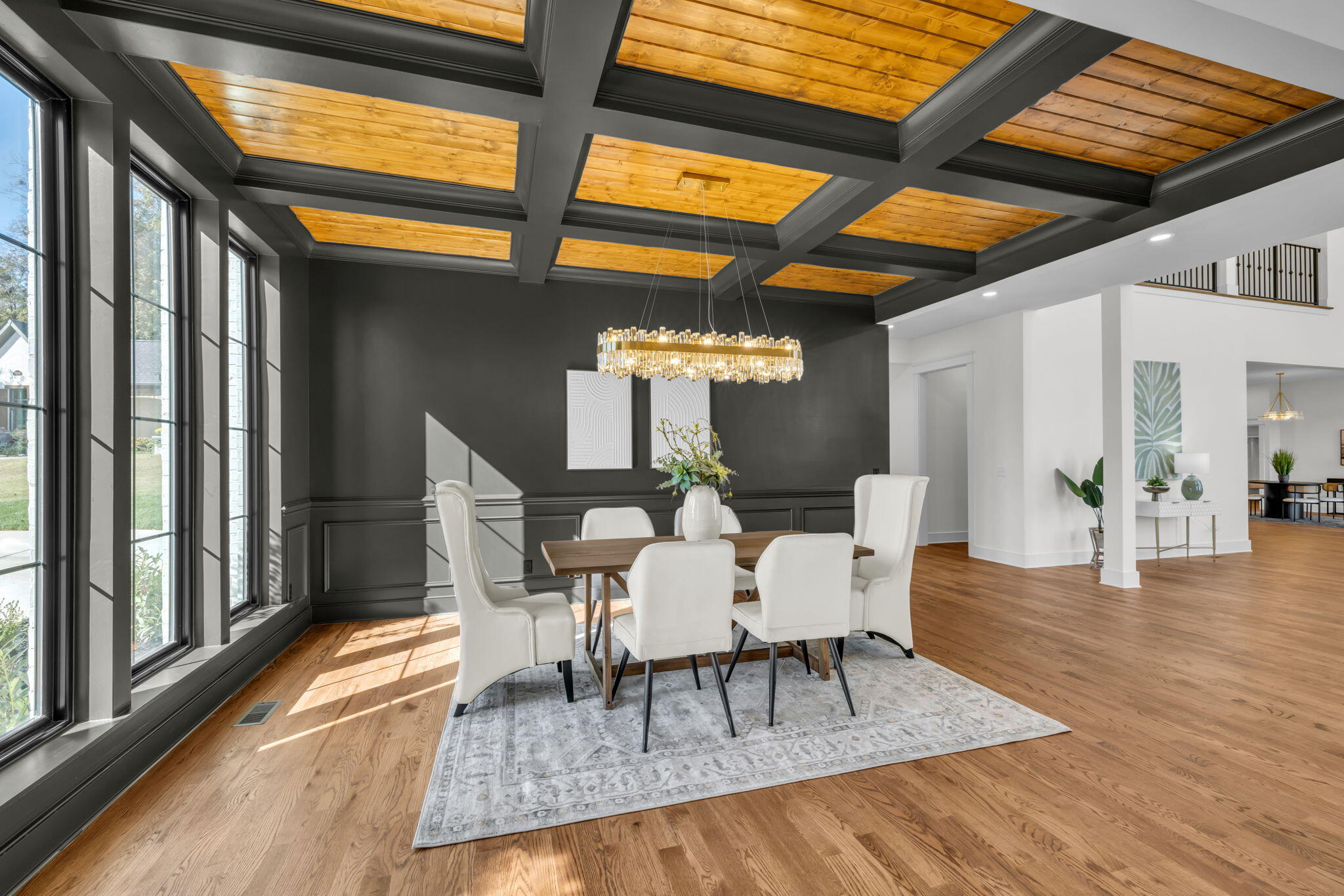 Dining room with white chairs, tall windows, and a wood-paneled coffered ceiling.