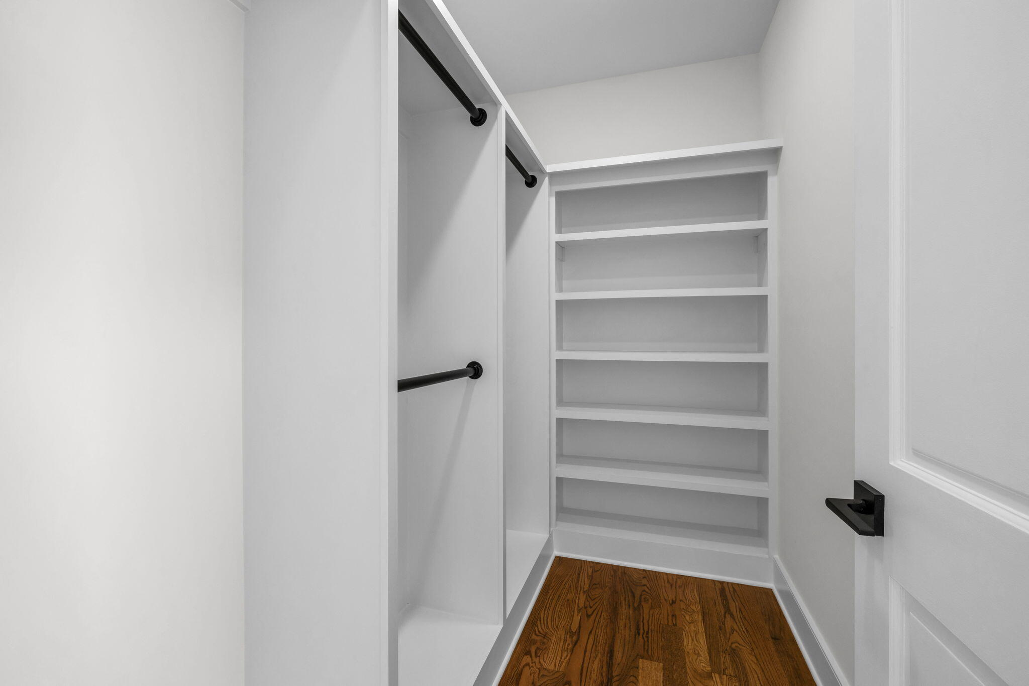 Walk-in closet with built-in white shelves, black hanging rods, and hardwood flooring.