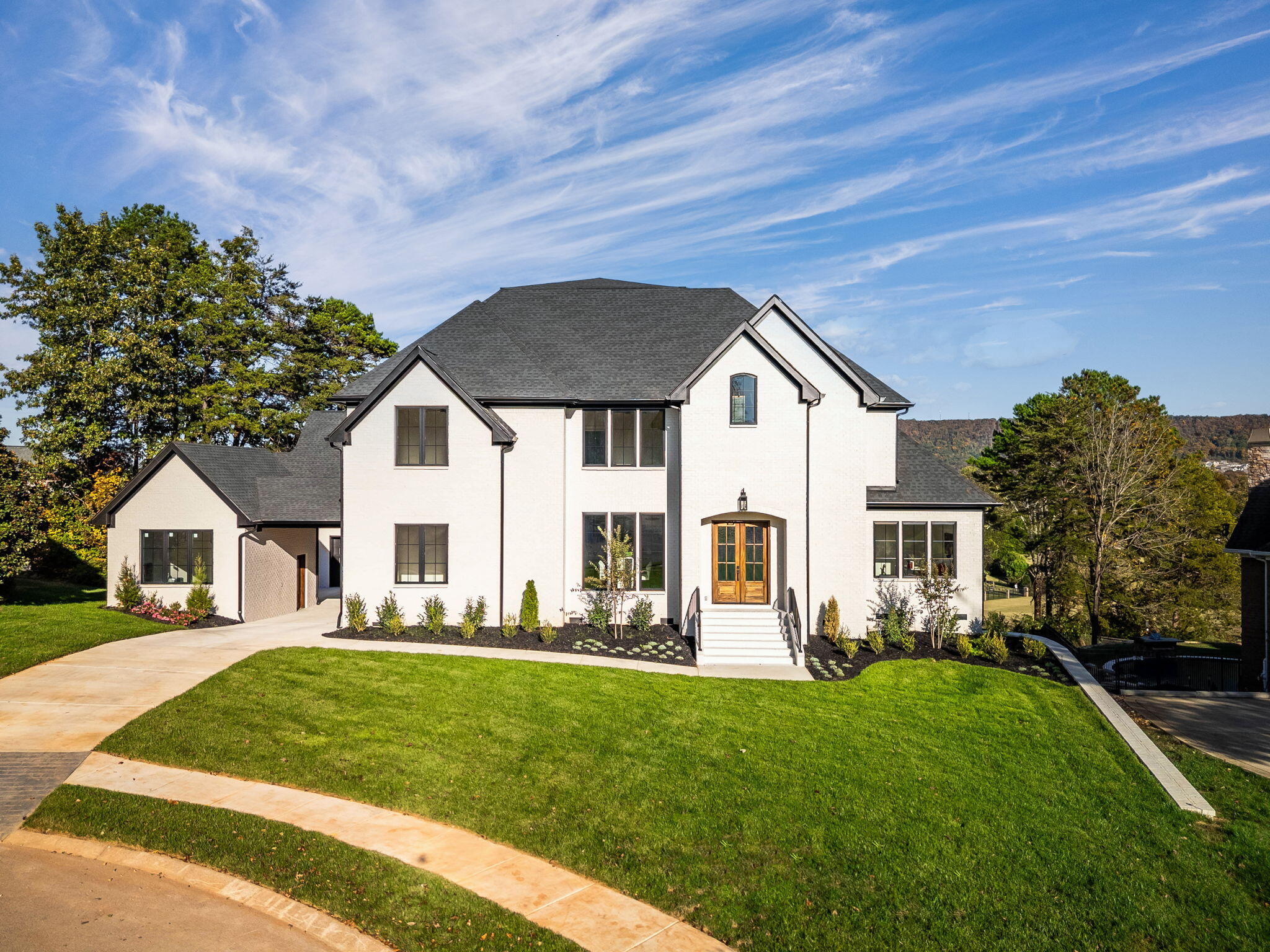 White two-story home with dark gabled roofs, wood entry doors, landscaped beds, and a broad front lawn.