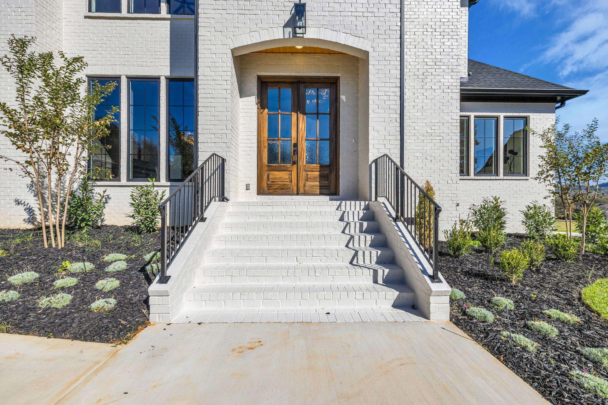 White brick home entrance with broad steps, black railings, and double wood-and-glass doors.