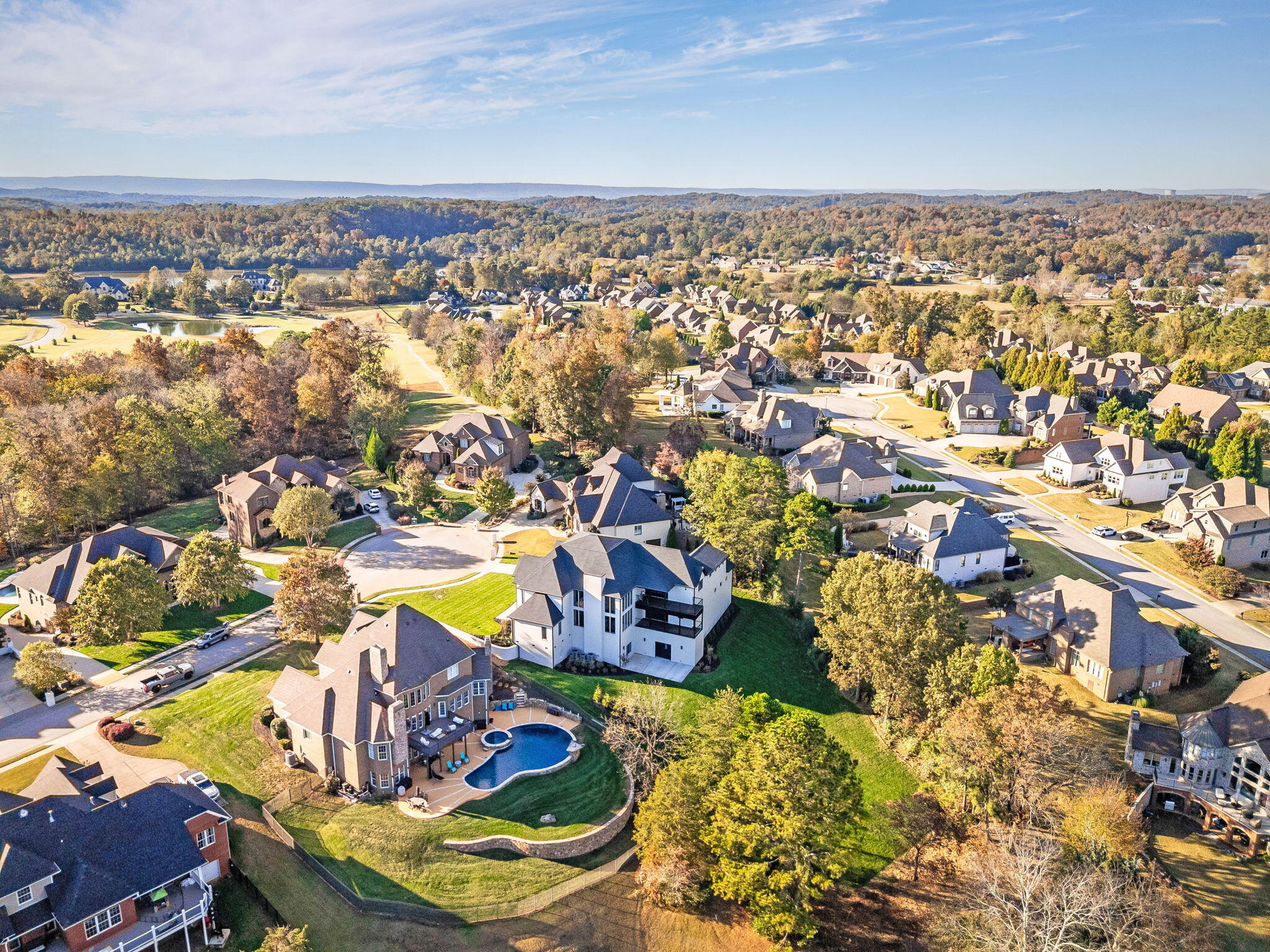 Aerial view of a residential neighborhood with a backyard pool, small pond, and wooded hills.