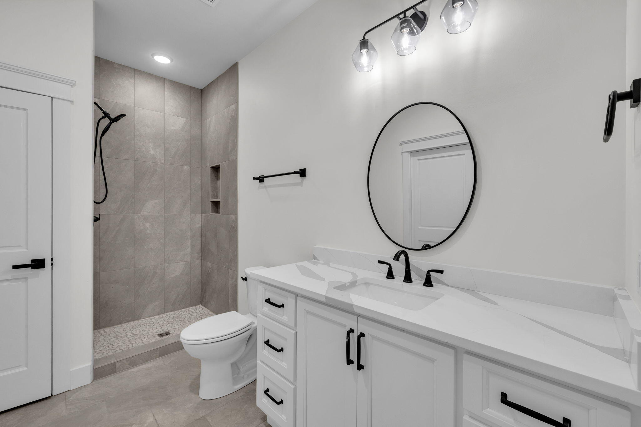 Modern bathroom with a round mirror, white vanity, black fixtures, and a tiled walk-in shower.