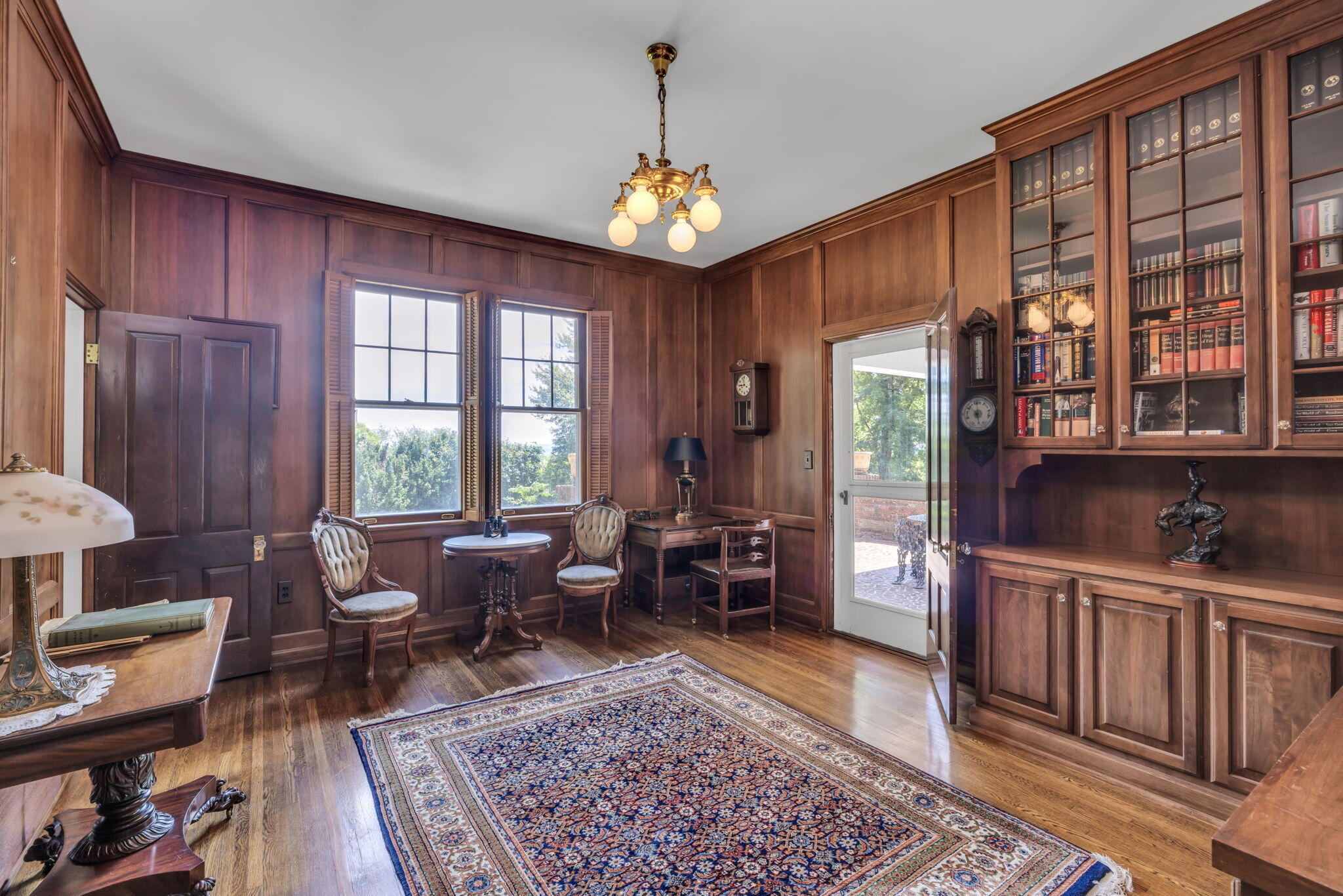 Wood-paneled study with built-in bookcases, antique seating, tall windows, and a patterned rug.