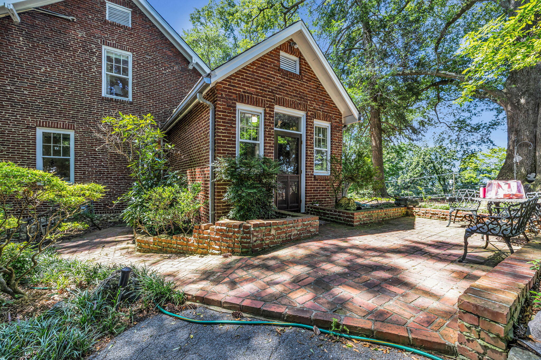 Red-brick house and broad brick patio shaded by mature trees, with raised planters and metal seating.