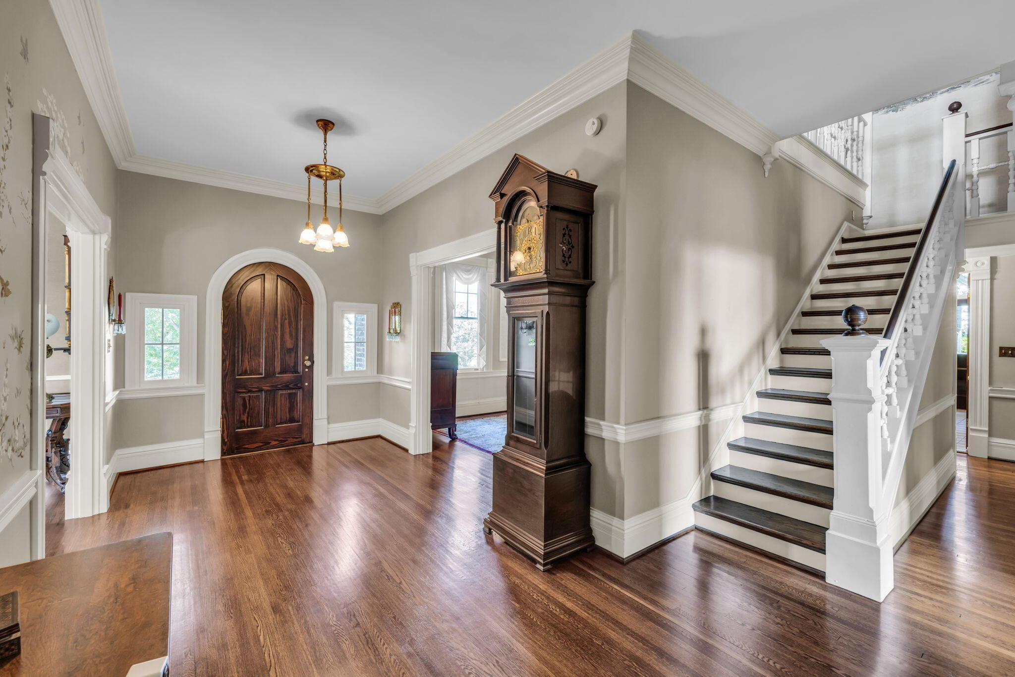 Entry hall with a dark arched front door, grandfather clock, and white staircase with dark treads.