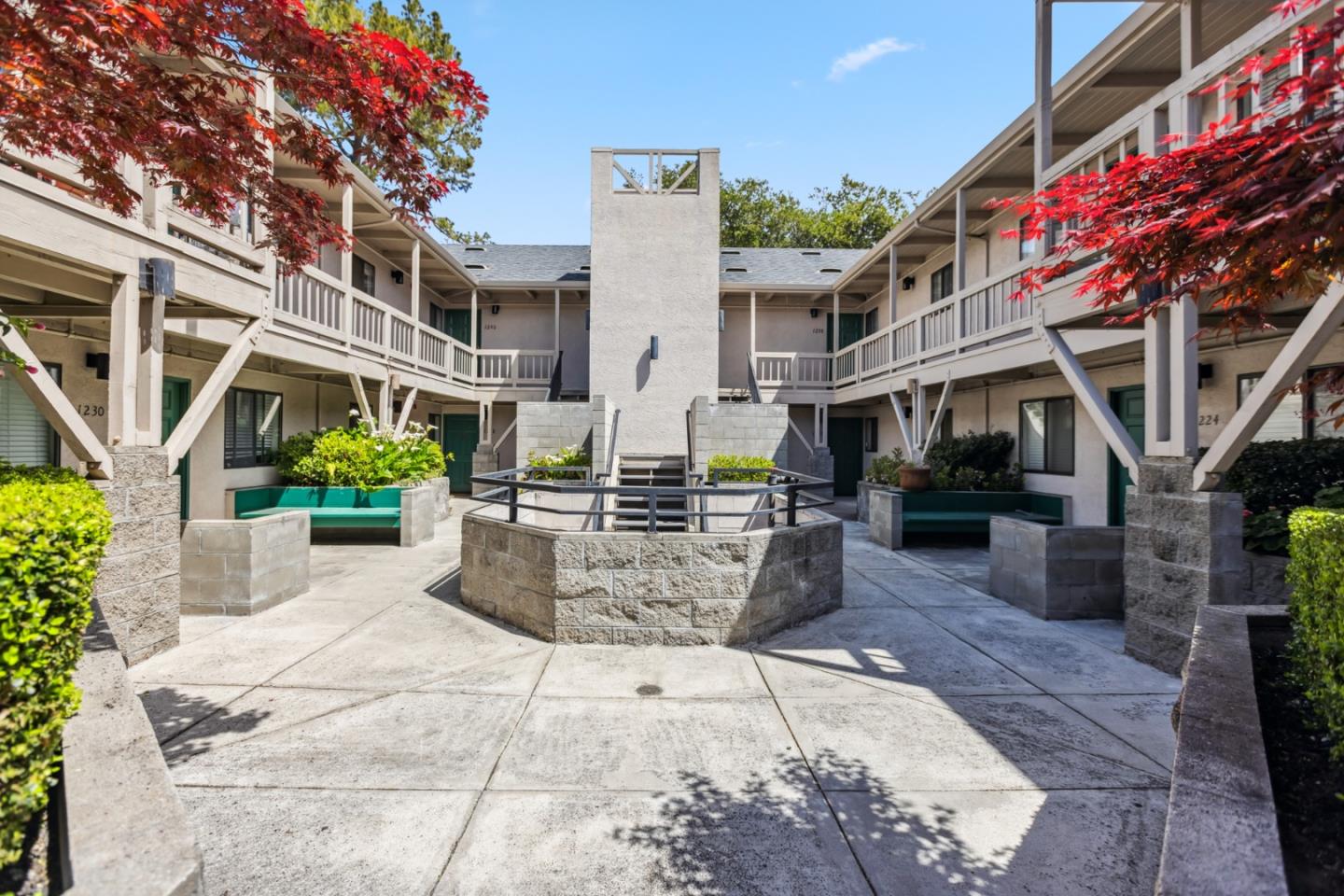 Apartment building courtyard with a raised stone planter, teal benches, and red-leaved trees.
