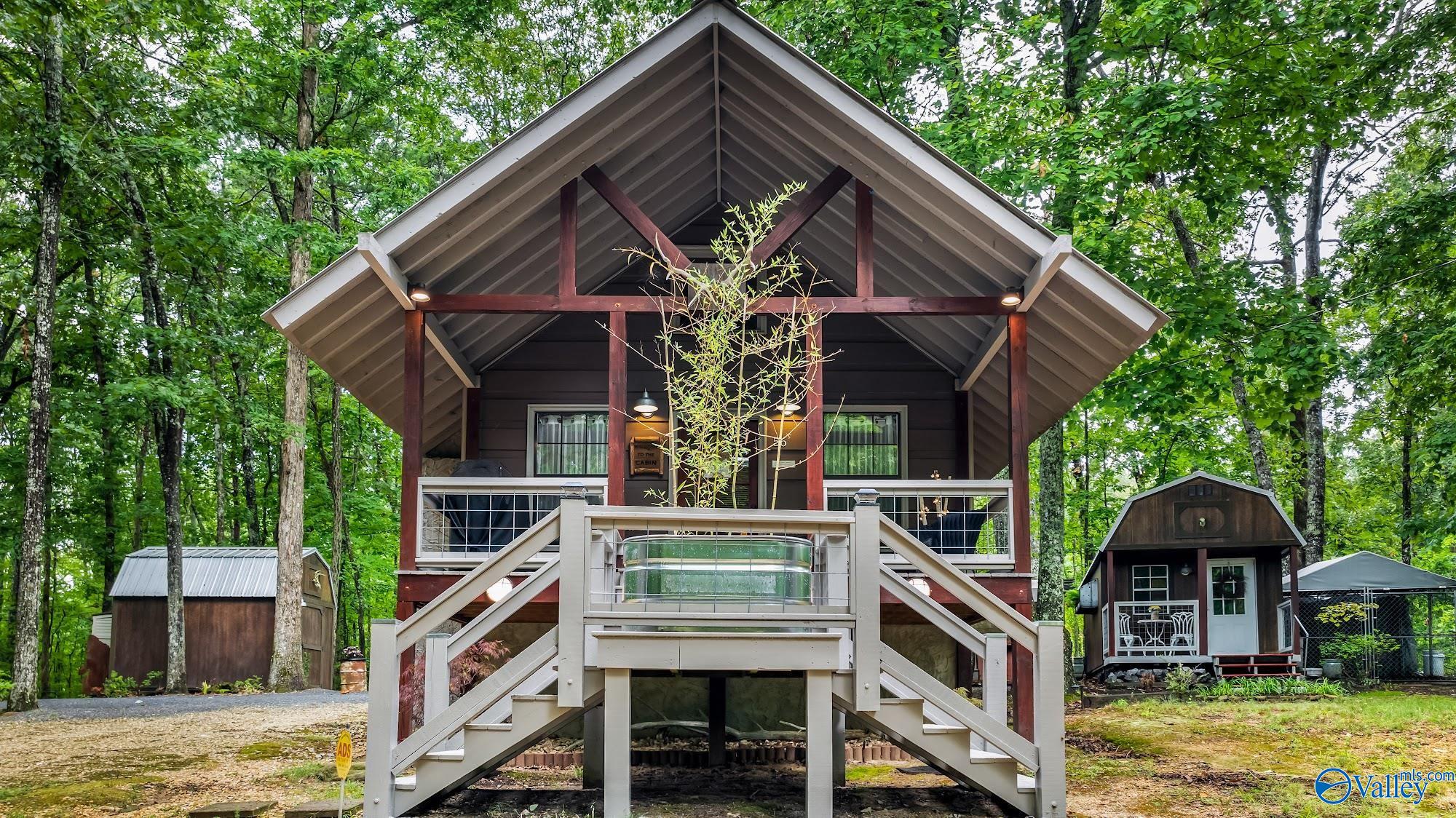 Raised cabin with a steep gabled roof and broad front stairs, surrounded by trees and outbuildings.