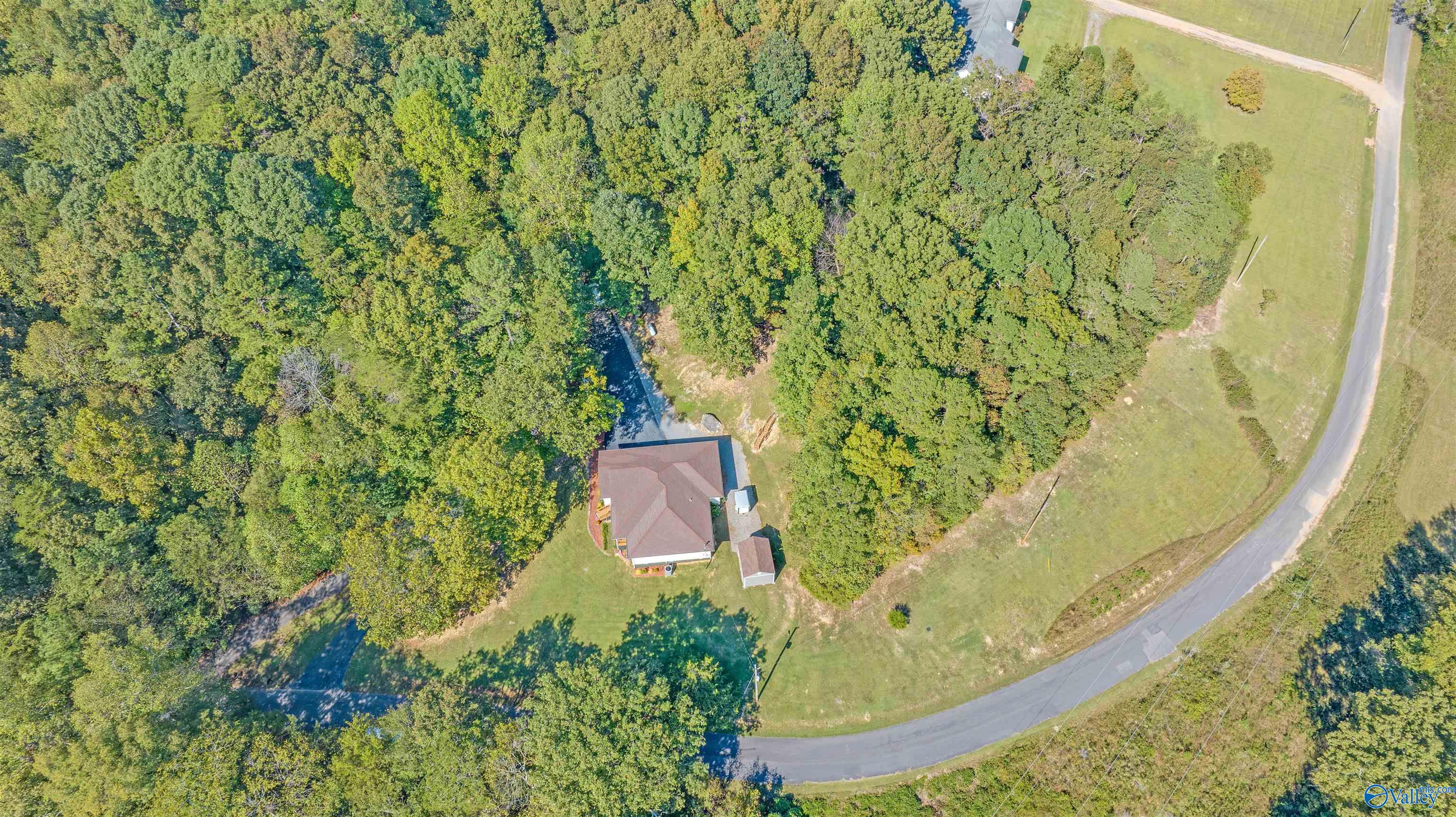 Aerial view of a house and outbuilding amid dense trees beside a curved road.