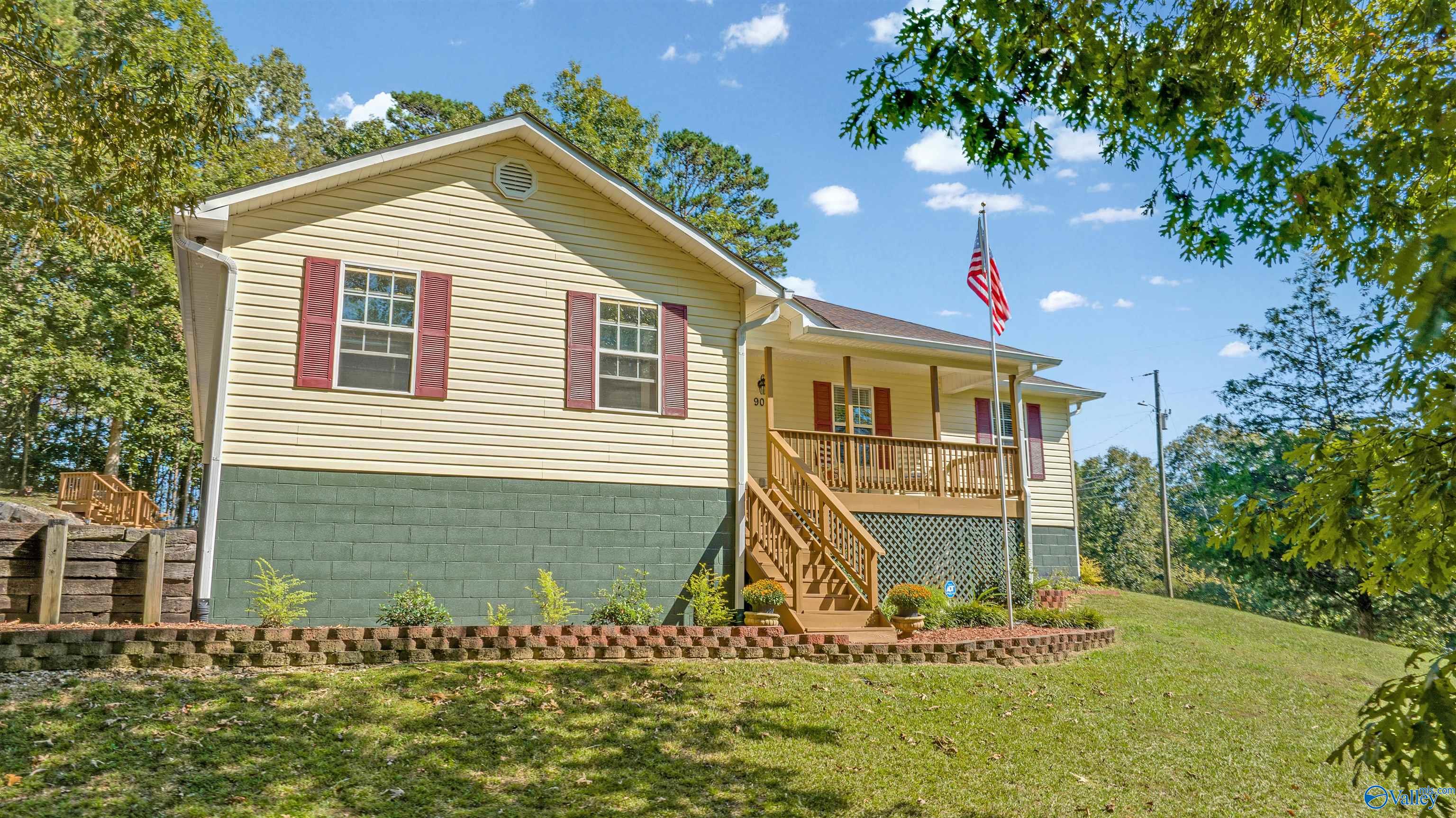 Yellow home with red shutters, raised wooden porch, American flag, and landscaped lawn.